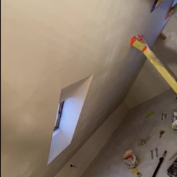 Usługi ogólnobudowlane - Wnętrze po gładziowaniu ścian i sufitu, widoczne okno w skosie, narzędzia malarskie na podłodze. Ściany w jasnym odcieniu beżu, gotowe do malowania.