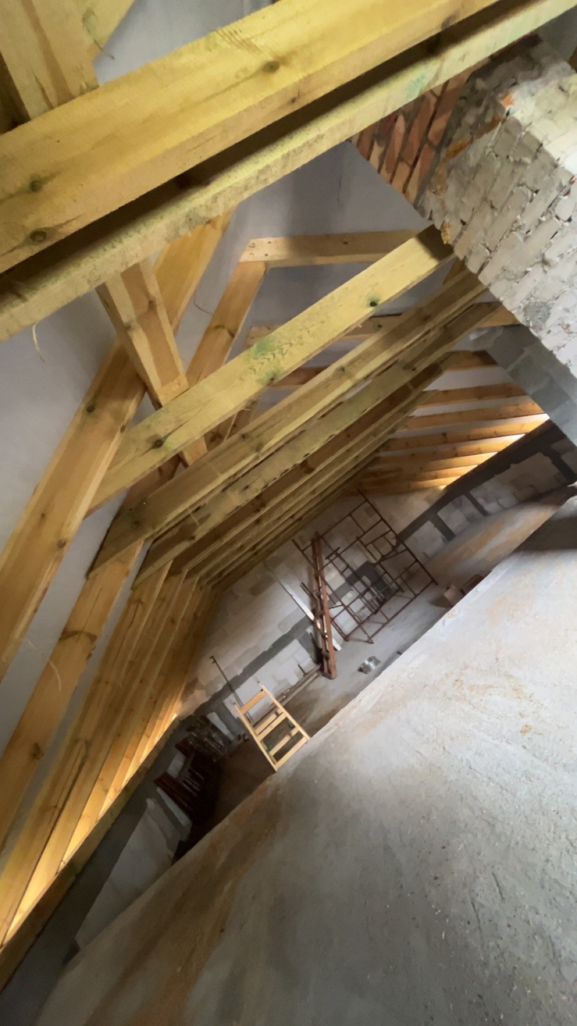 Konstrukcja drewniana dachu poddasza z widocznym kominem z cegły, drabina i rusztowanie w tle. Widok od dołu, oświetlenie naturalne.