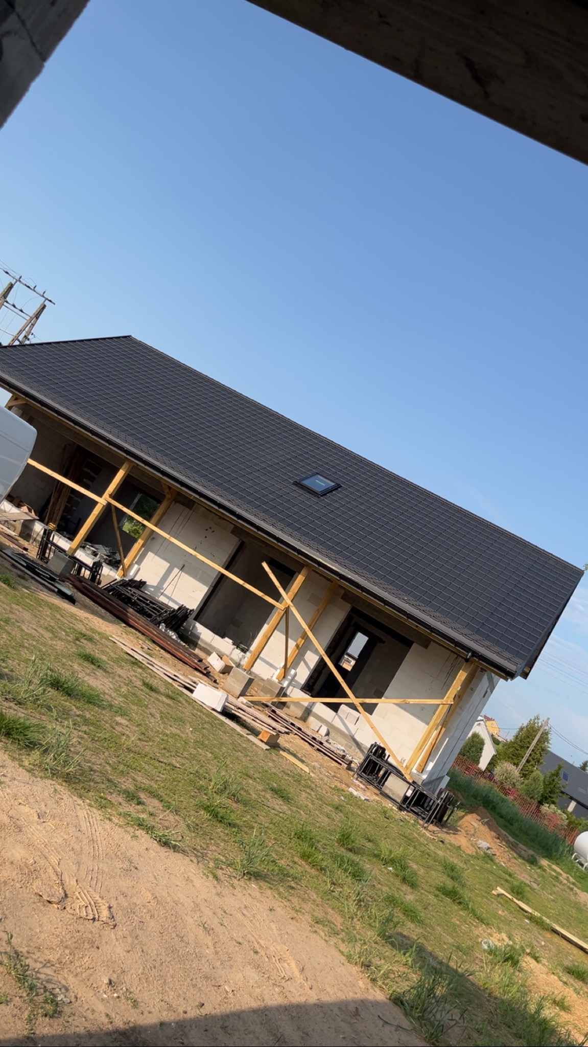 Dom w trakcie budowy z czarnym dachem i oknem dachowym, widok z boku, drewniane podpory, częściowo otynkowane ściany, w tle błękitne niebo.