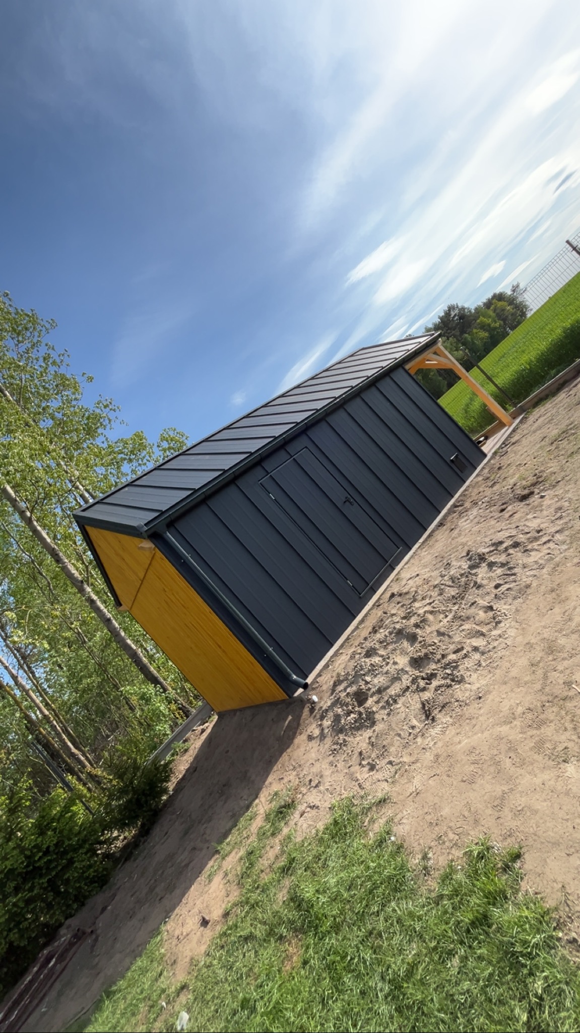 Gotowy, nowoczesny domek narzędziowy z czarną blachą i żółtymi detalami, usytuowany na trawiastym terenie pod błękitnym niebem. Widoczne drzewa i pola w tle.