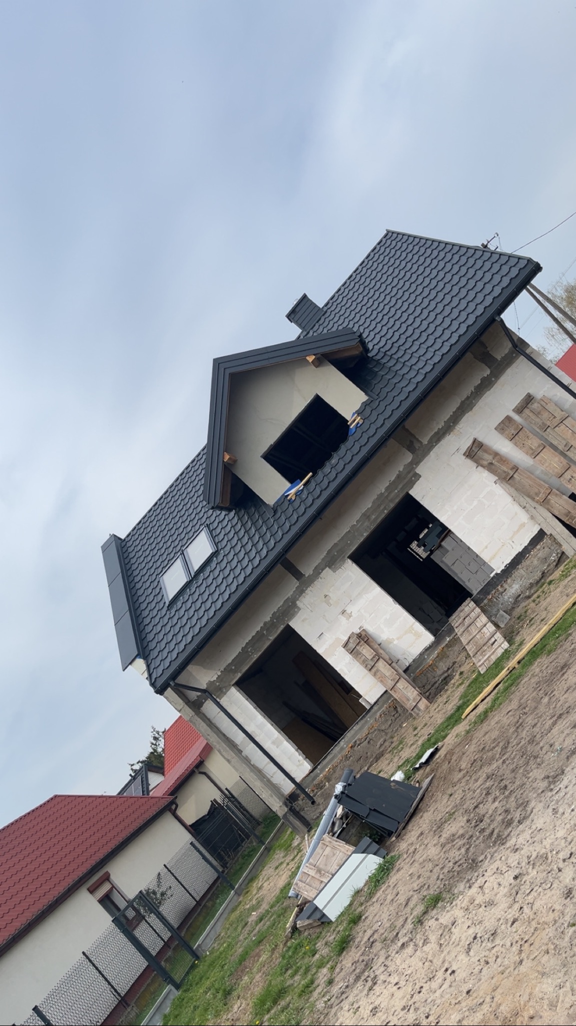 Dom w stanie surowym z czarną dachówką, widoczne elementy konstrukcyjne i okna dachowe. Ujęcie z dołu, podkreślające geometrię dachu i bryłę budynku.