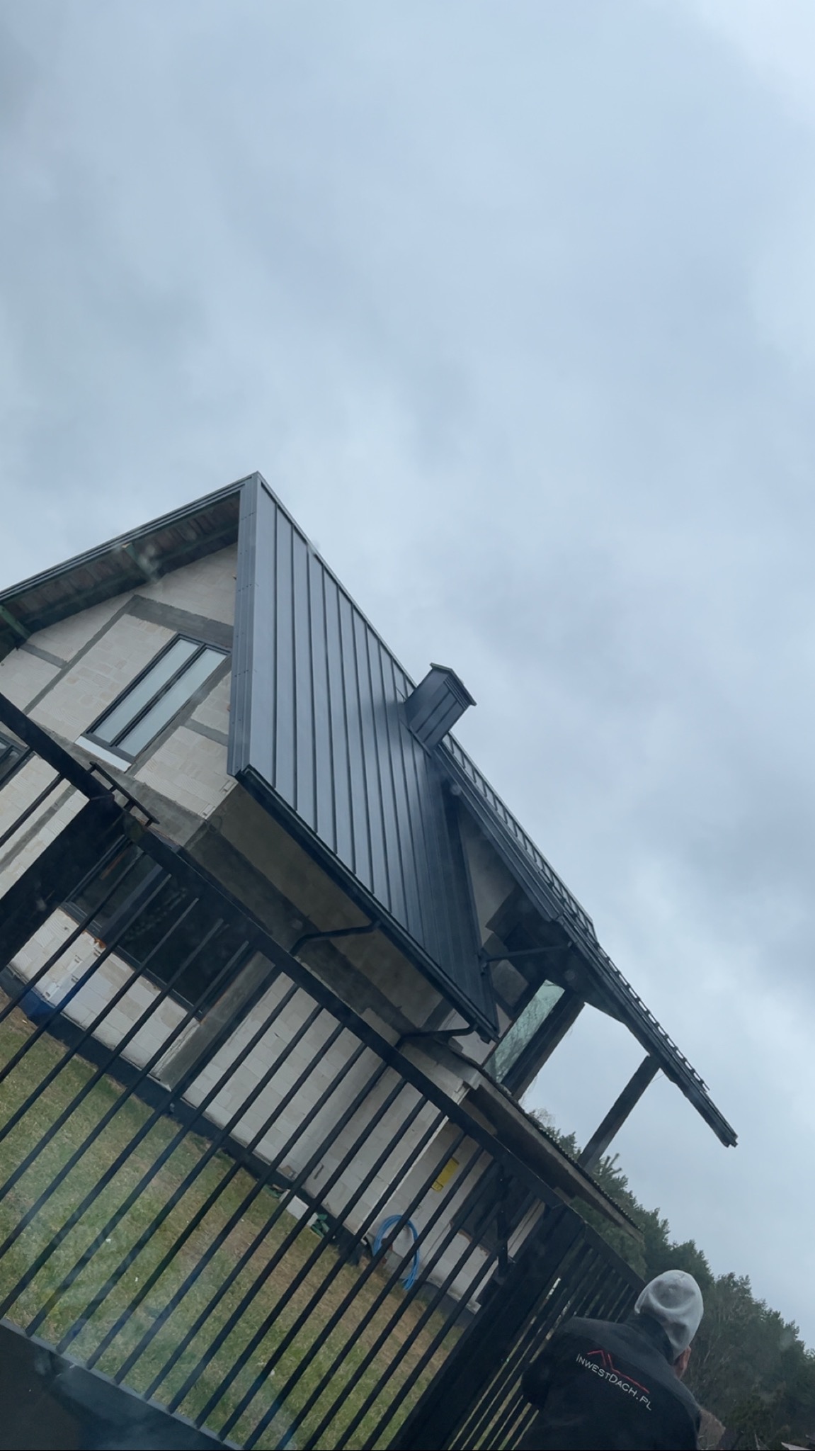 Nowoczesny dom z grafitowym dachem z blachy na rąbek stojący, widoczny zza czarnego ogrodzenia. Pochmurne niebo w tle, fragment sylwetki osoby w prawym dolnym rogu.