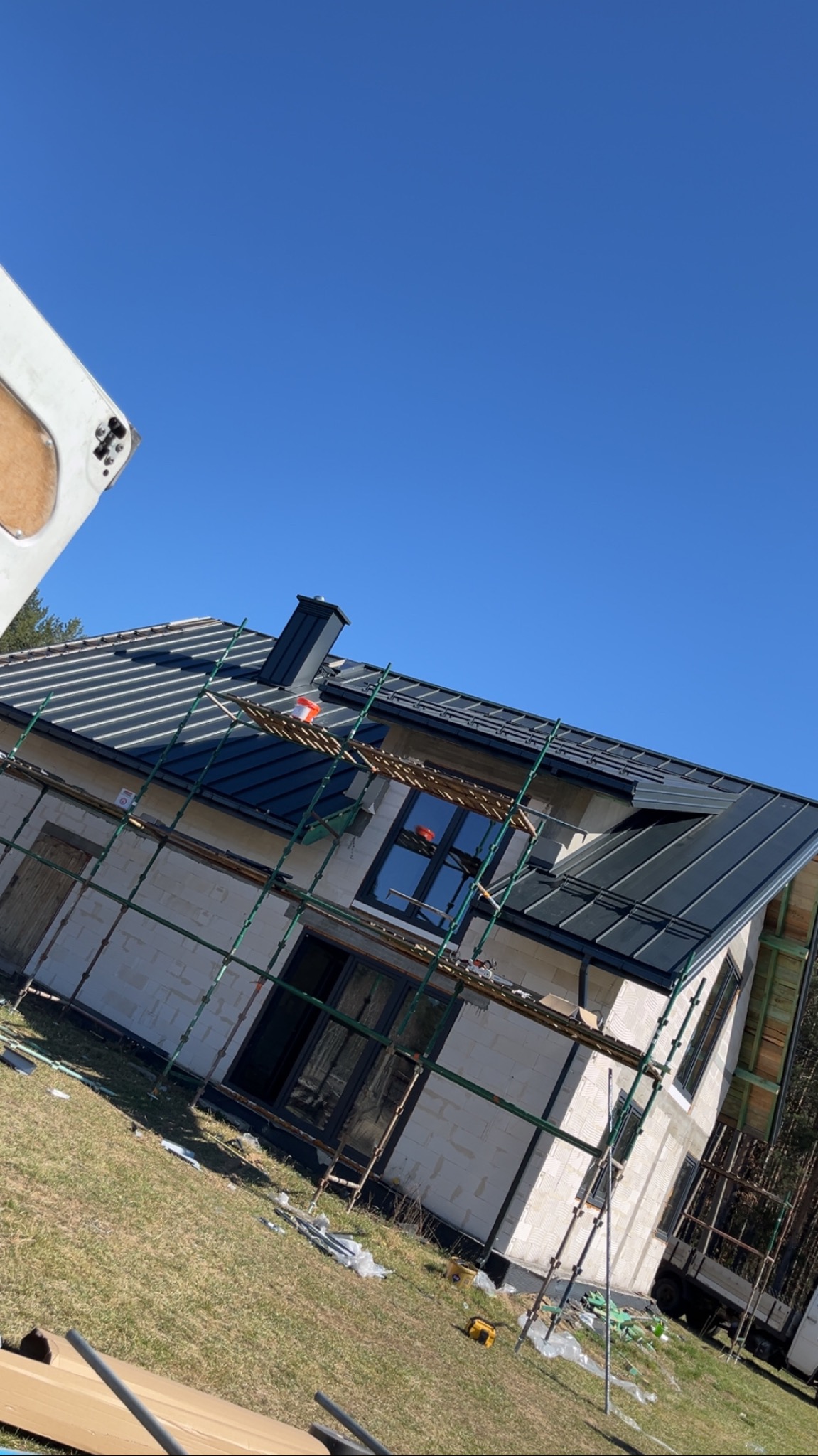 Dom w trakcie budowy z ciemnym dachem z blachy, z widocznymi rusztowaniami i elementami konstrukcyjnymi, na tle błękitnego nieba.