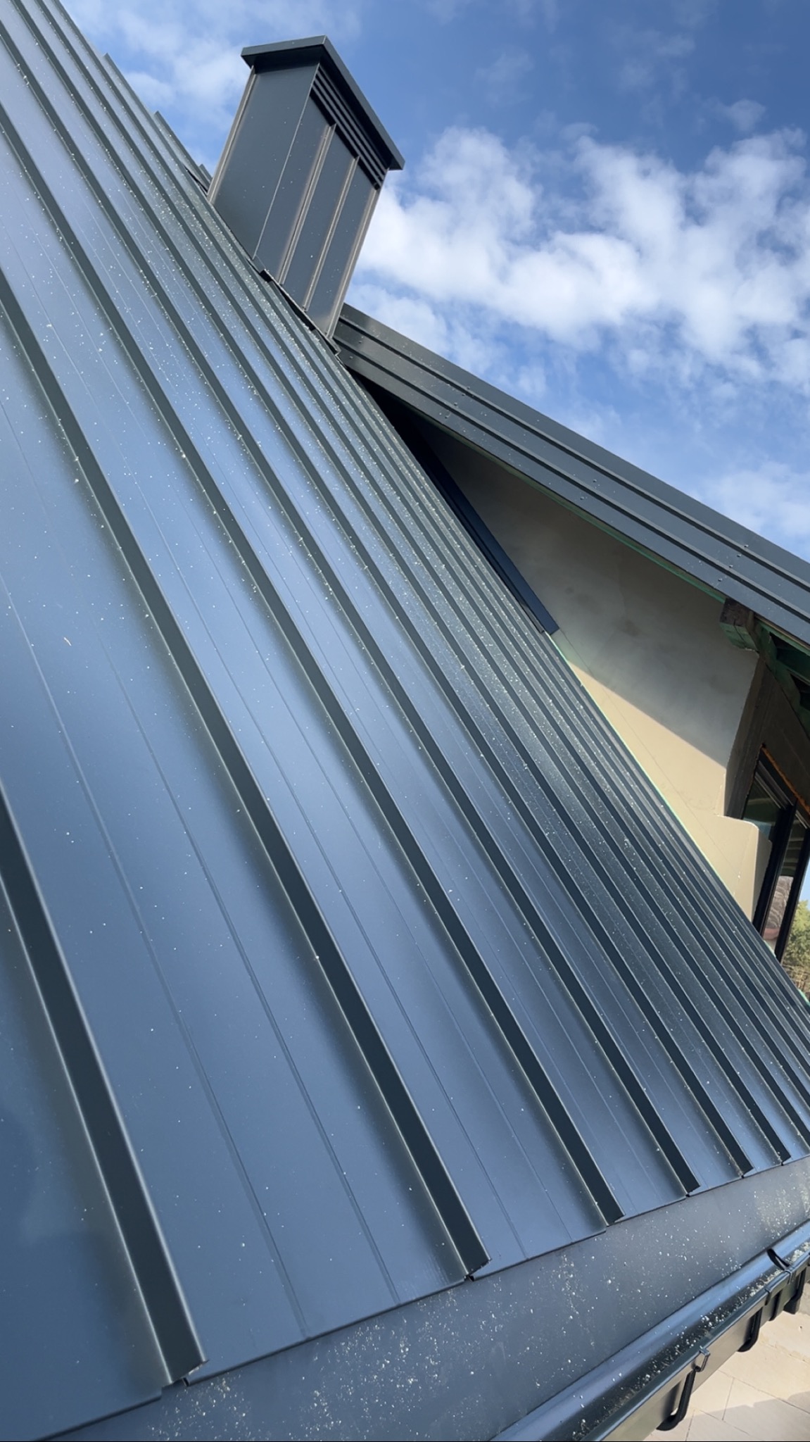 Szary dach z blachy trapezowej, widok pod kątem. Na dachu komin wentylacyjny w kolorze dachu. W tle błękitne niebo z chmurami.