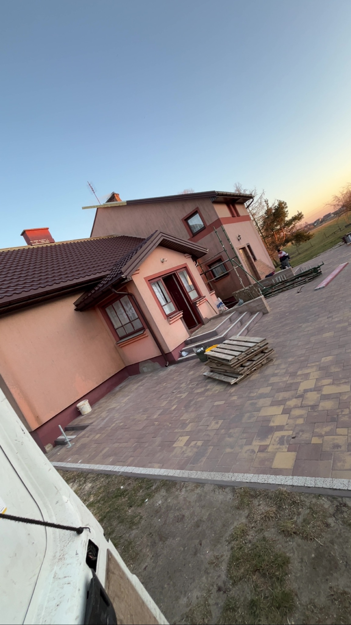 Ukończony dom z brązowym dachem i beżową elewacją, widok z góry. Na podwórku palety i rusztowania. Część białego samochodu w lewym dolnym rogu.