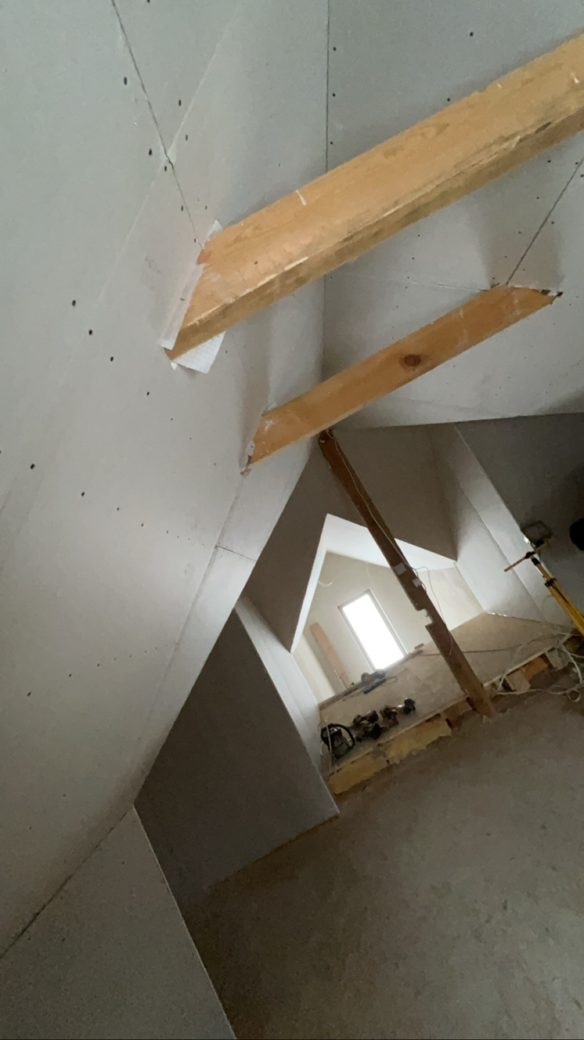 Skośne poddasze z zamontowaną płytą karton-gips, widoczne drewniane belki konstrukcyjne i fragment okna. Prace wykończeniowe w trakcie realizacji.