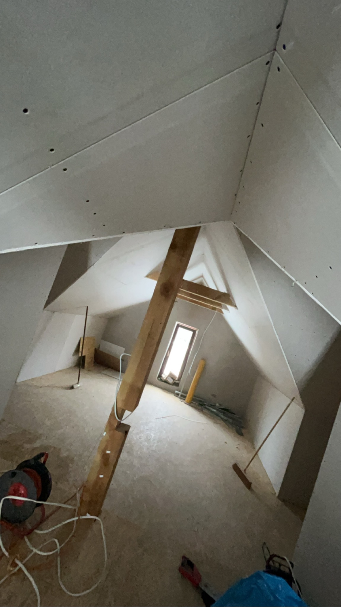 Poddasze z płytami karton-gips, widoczne drewniane elementy konstrukcyjne, narzędzia i okno. Prace wykończeniowe w trakcie realizacji.