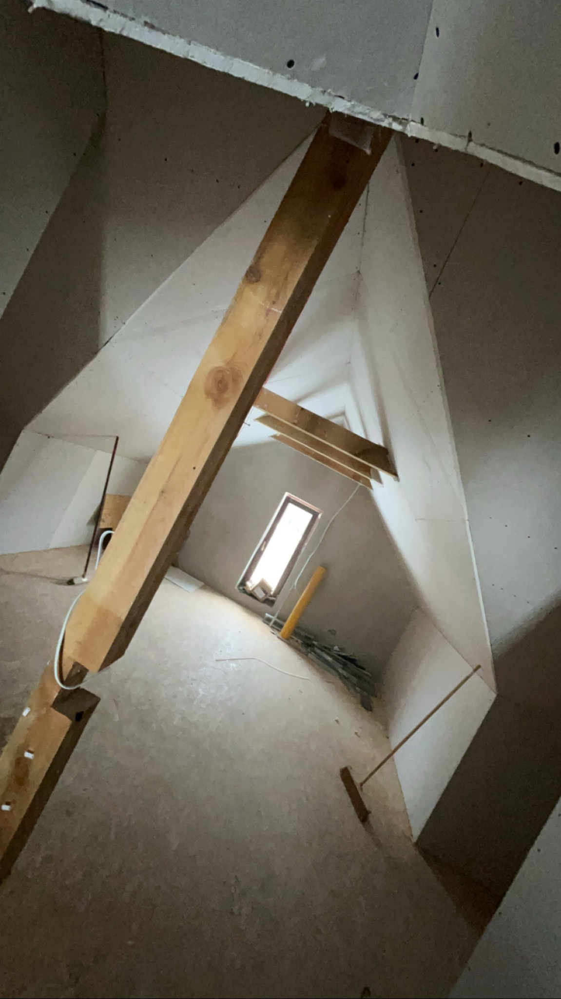 Poddasze w trakcie prac wykończeniowych: drewniane belki, surowe ściany z płyt kartonowo-gipsowych, okno w tle. Widoczne narzędzia i materiały budowlane.