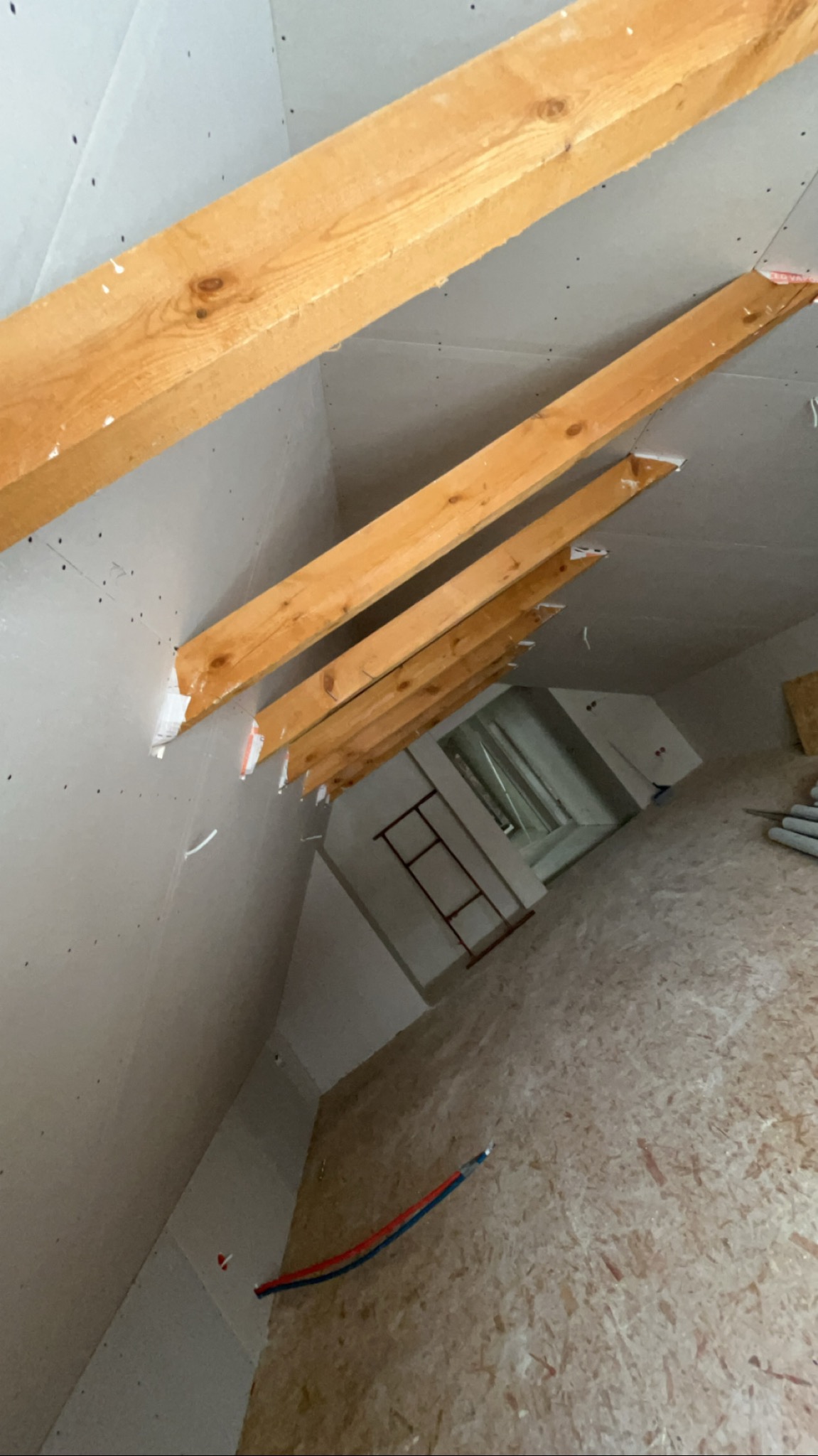 Poddasze w trakcie adaptacji: widoczne drewniane krokwie, płyty karton-gips na ścianach i skośny sufit. Na podłodze płyta OSB, w tle drabina i rury.