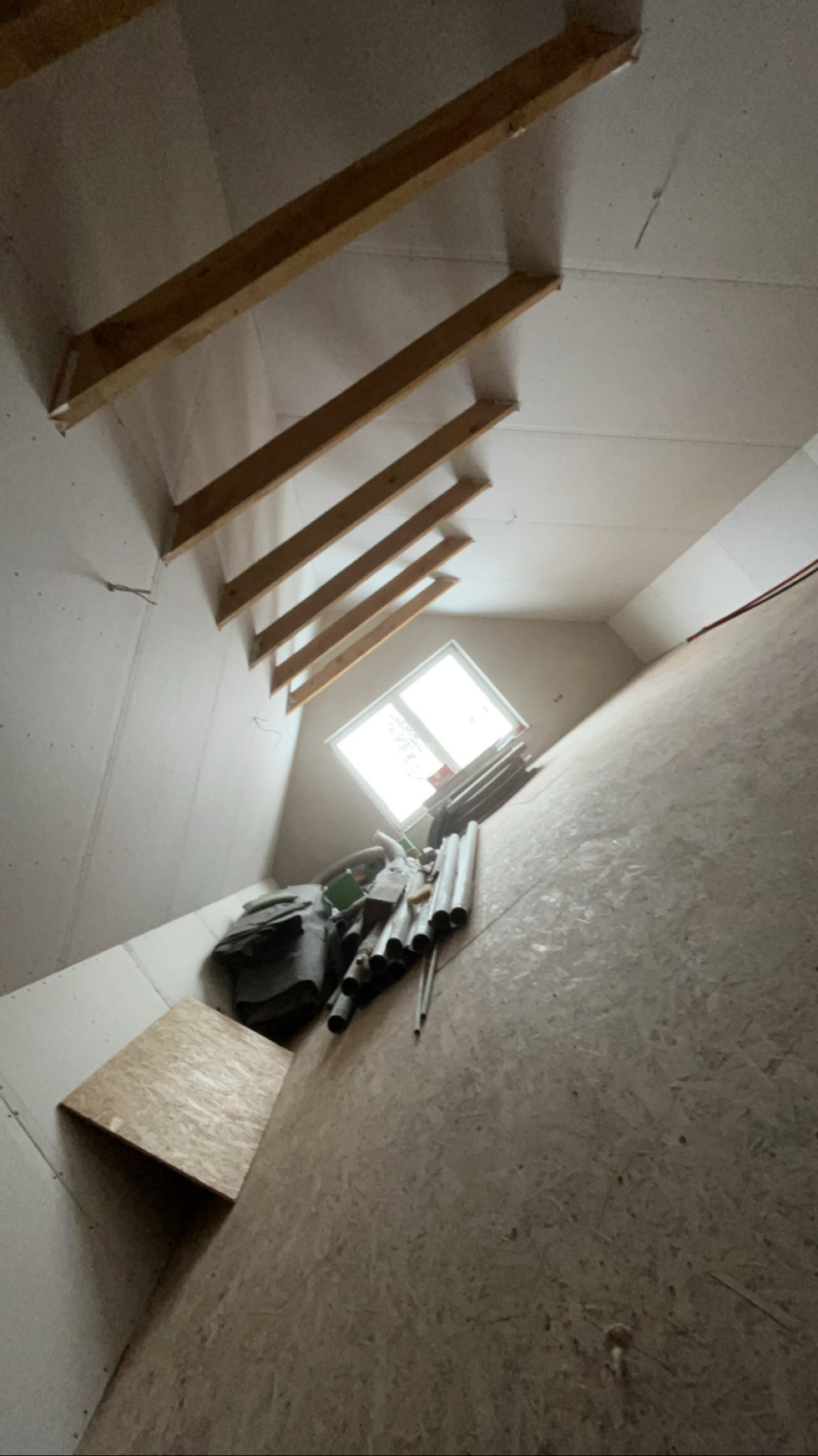 Poddasze w trakcie adaptacji: widoczne drewniane belki stropowe, ściany wyłożone płytami OSB, okno dachowe i materiały budowlane. Skos dachu, surowy stan wykończenia.