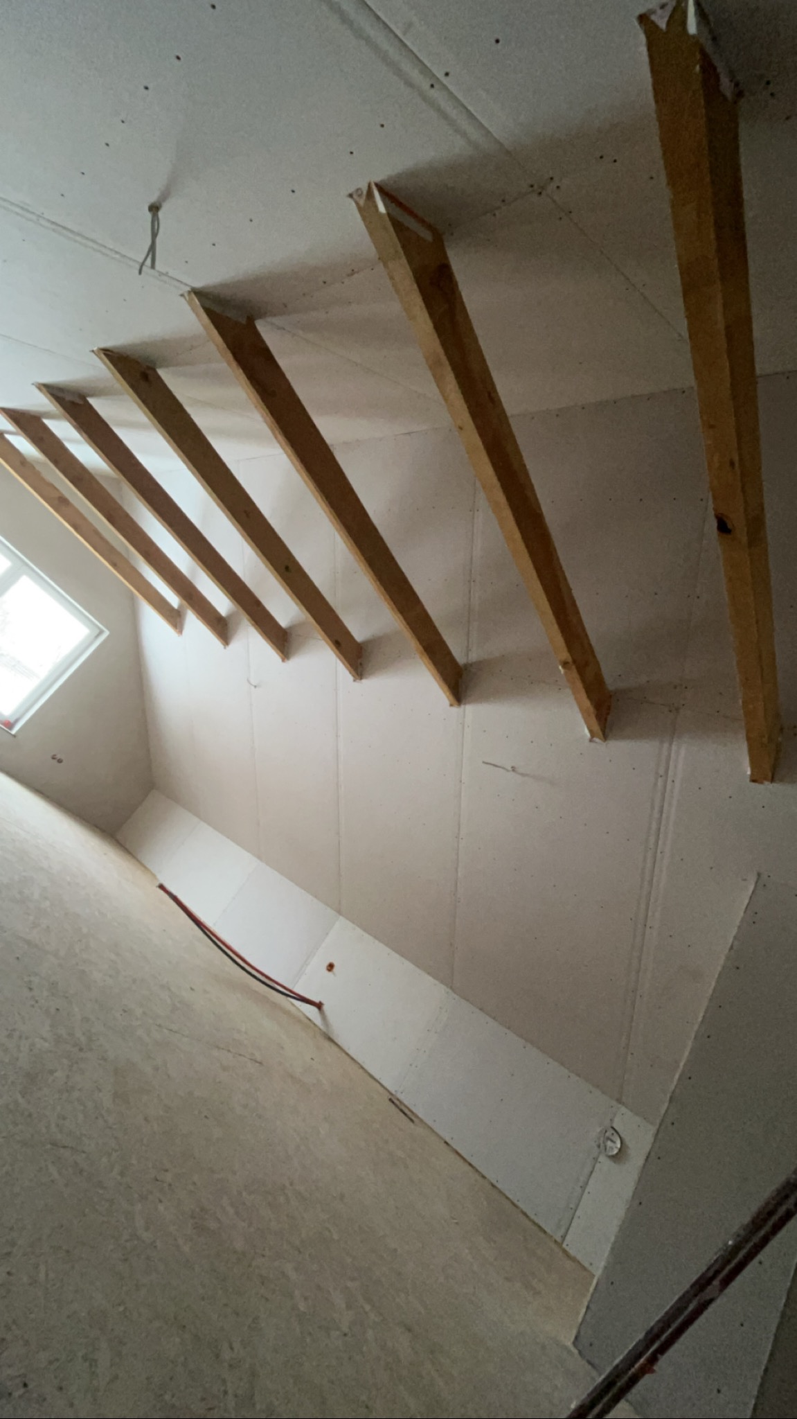 Skośne poddasze z drewnianą konstrukcją i płytami kartonowo-gipsowymi. Widoczne kable i fragment okna. Ujęcie z dołu, w jasnej tonacji.