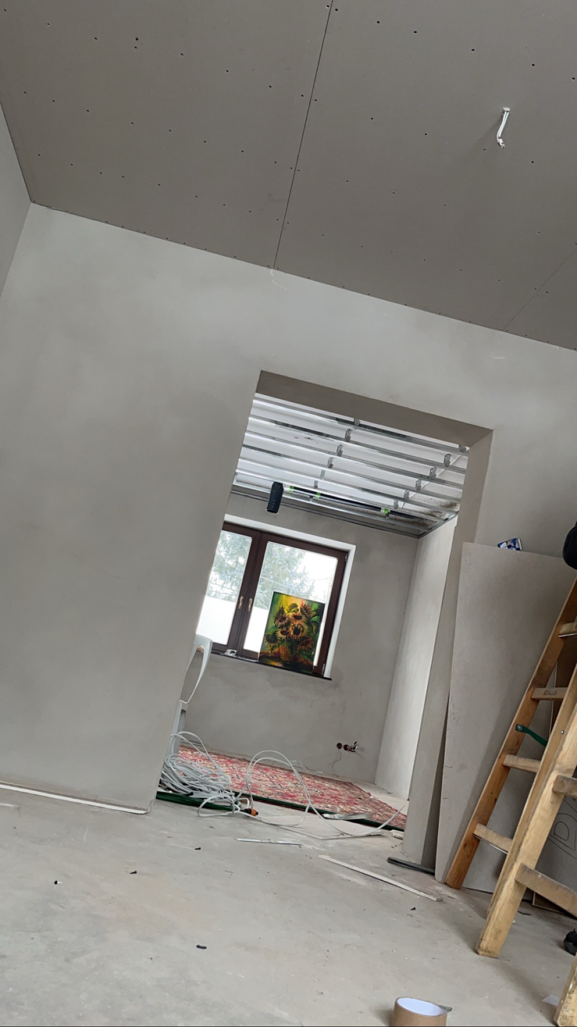 Wnętrze w trakcie prac: sufit z płyt karton-gips, widoczne profile montażowe. Na podłodze kable i dywan, drabina oparta o ścianę. W oknie obraz.