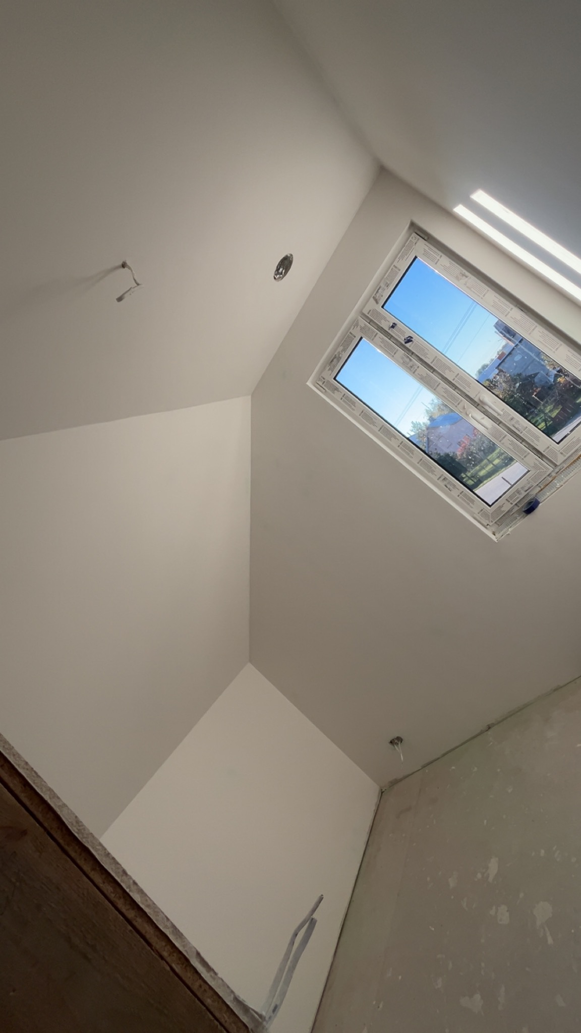 Poddasze po tynkowaniu na biało, widoczne okno dachowe z widokiem na zewnątrz i wystające kable. Ściany gładkie, przygotowane do dalszych prac wykończeniowych.
