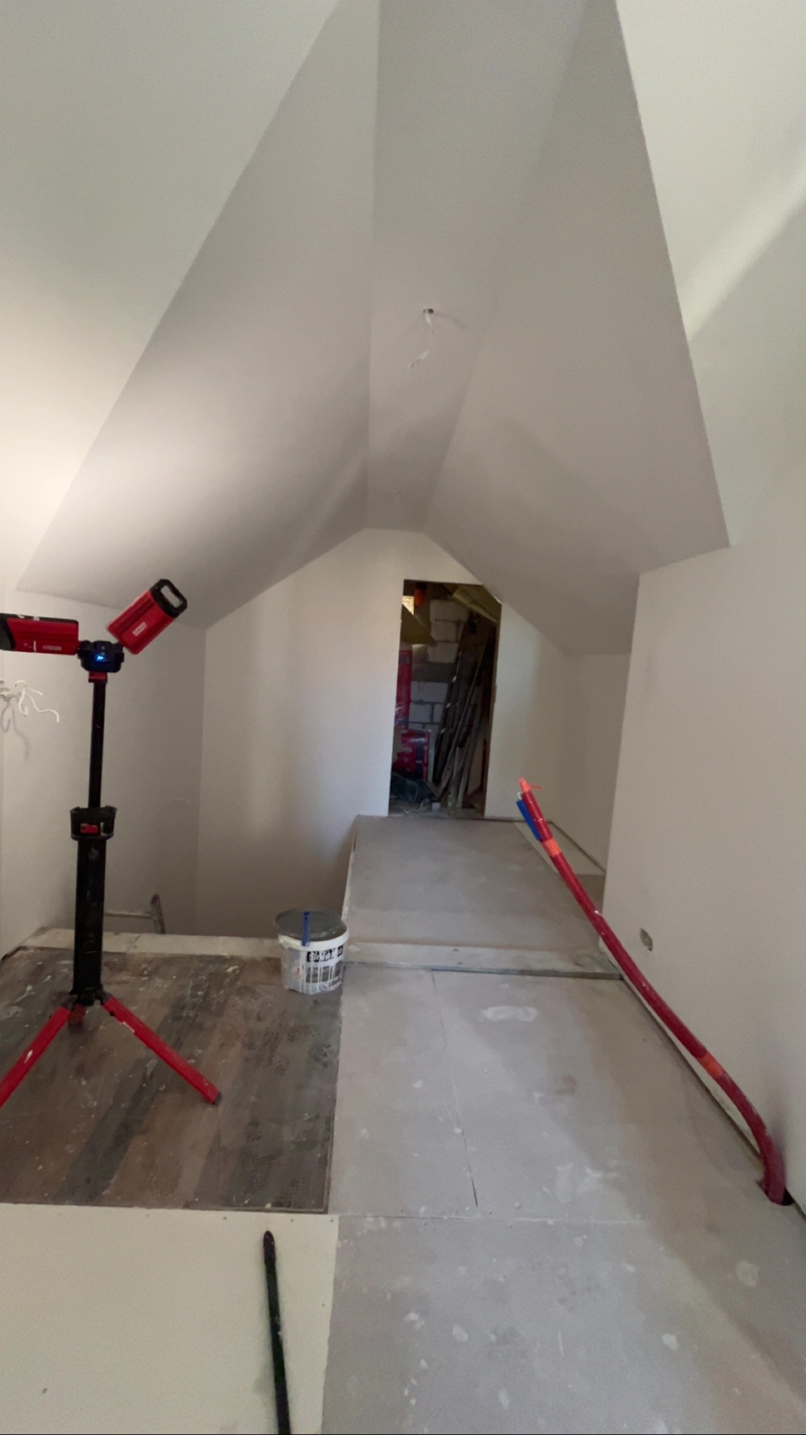 Poddasze w trakcie remontu: surowe ściany, widoczny stelaż, lampa budowlana na statywie, przygotowane podłoże. Prace wykończeniowe w toku.