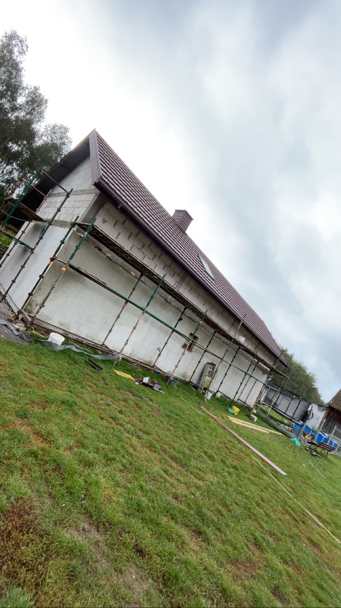 Budynek w trakcie ocieplania elewacji, widoczne rusztowania i częściowo otynkowana ściana. Dach z brązowej blachy, pochmurne niebo w tle. Ujęcie z dołu.