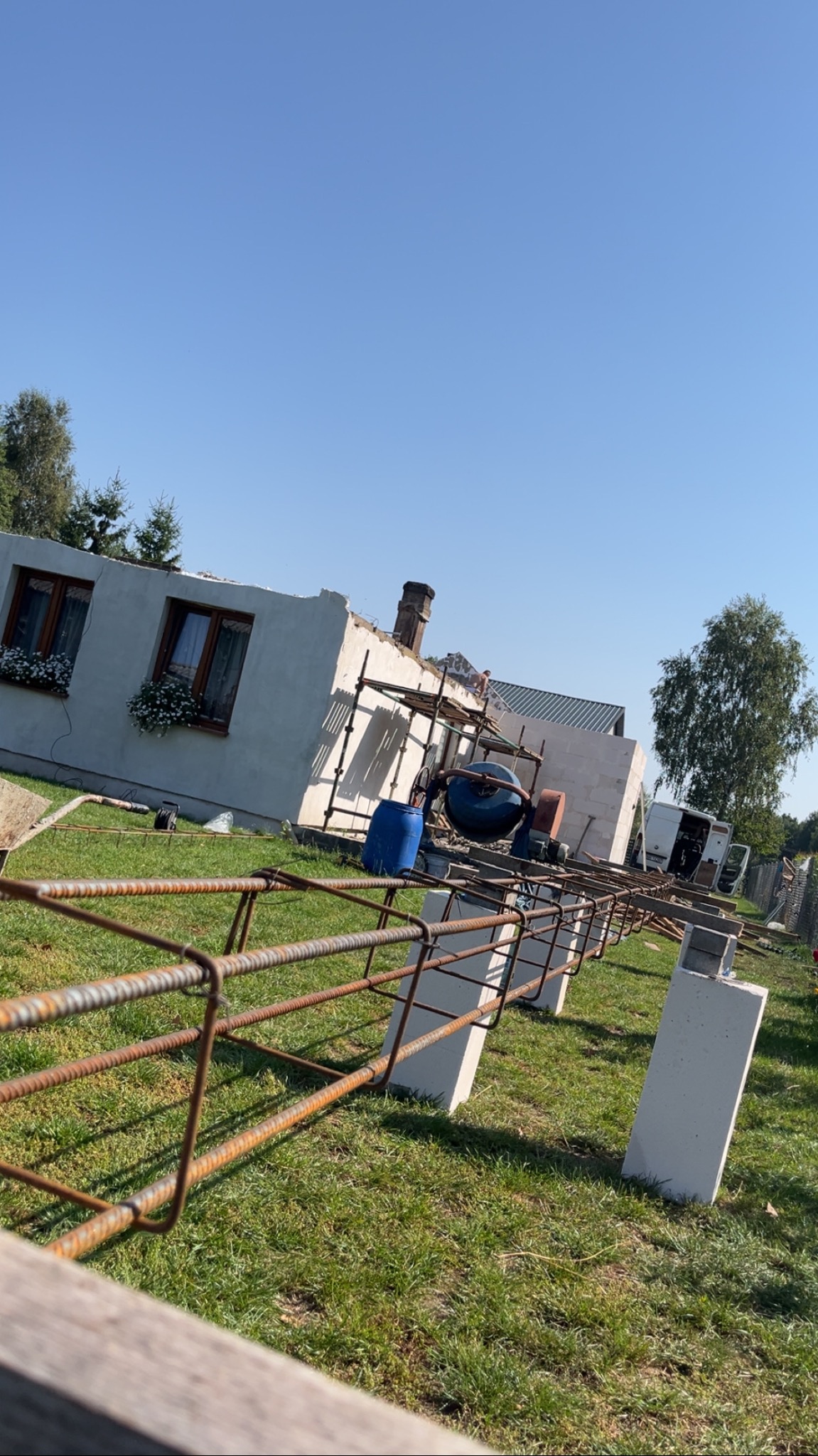 Budowa domu w toku: widoczne fundamenty, zbrojenie, ściany z pustaków i rusztowania. Na dachu pracownik. Betonarka i beczka na trawie. Słoneczny dzień.