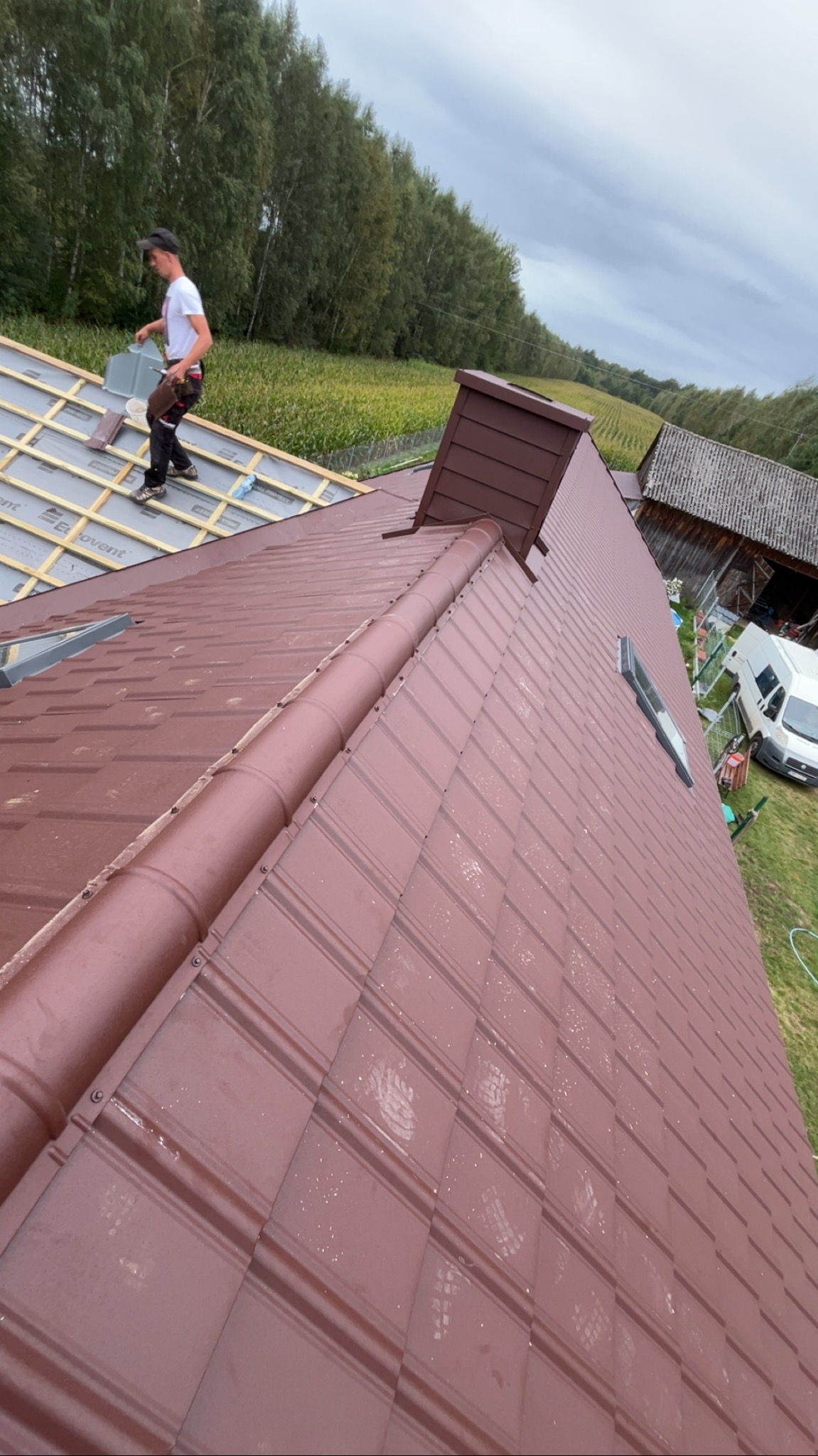 Praca dekarza w Zawistach podleśnych: brązowy dach z blachy na ukończeniu, widoczny komin i okno dachowe, dekarz w trakcie montażu.