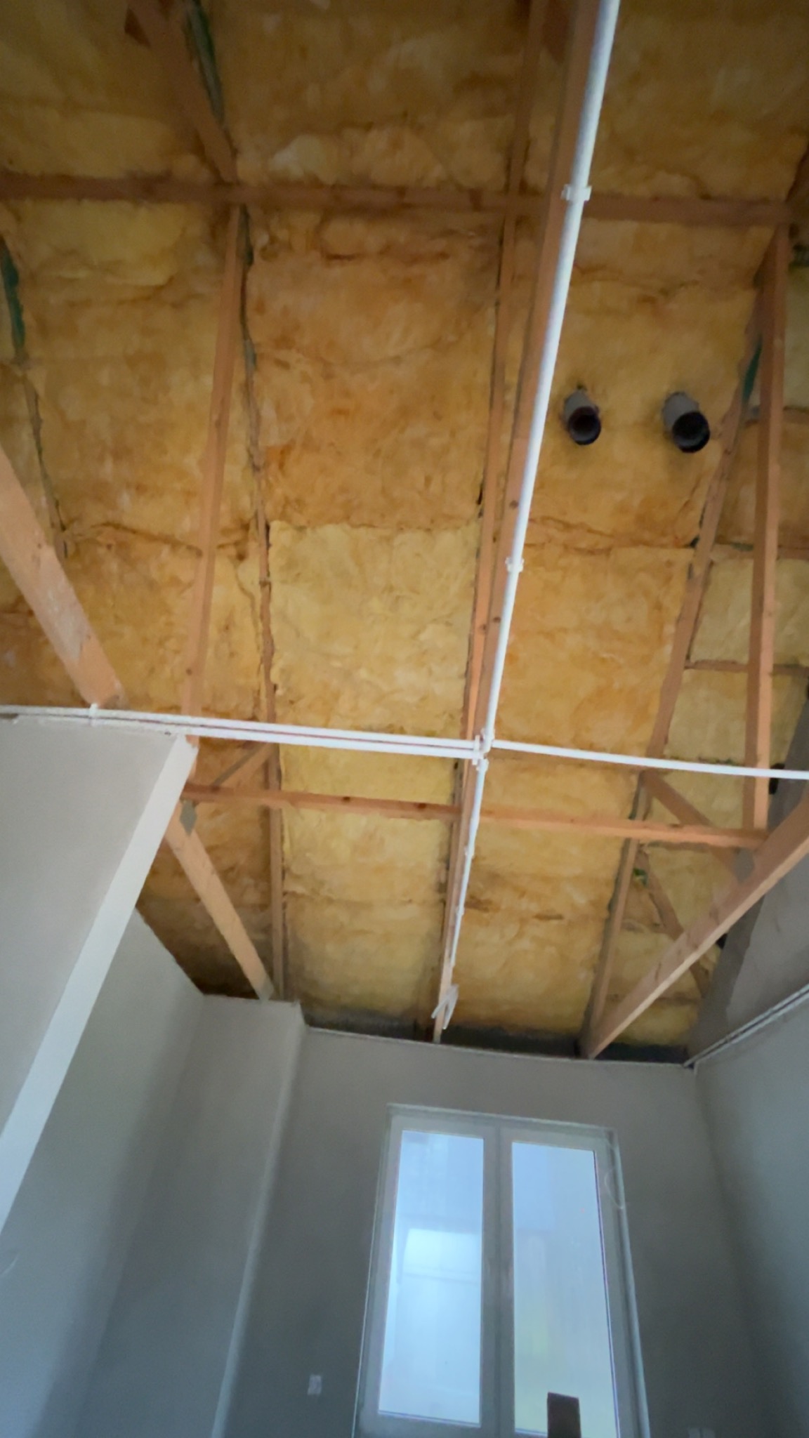 Poddasze z widoczną izolacją z wełny mineralnej między drewnianymi krokwiami. Widoczne rury instalacyjne i okno. Ujęcie z dołu, podkreślające strukturę izolacji.