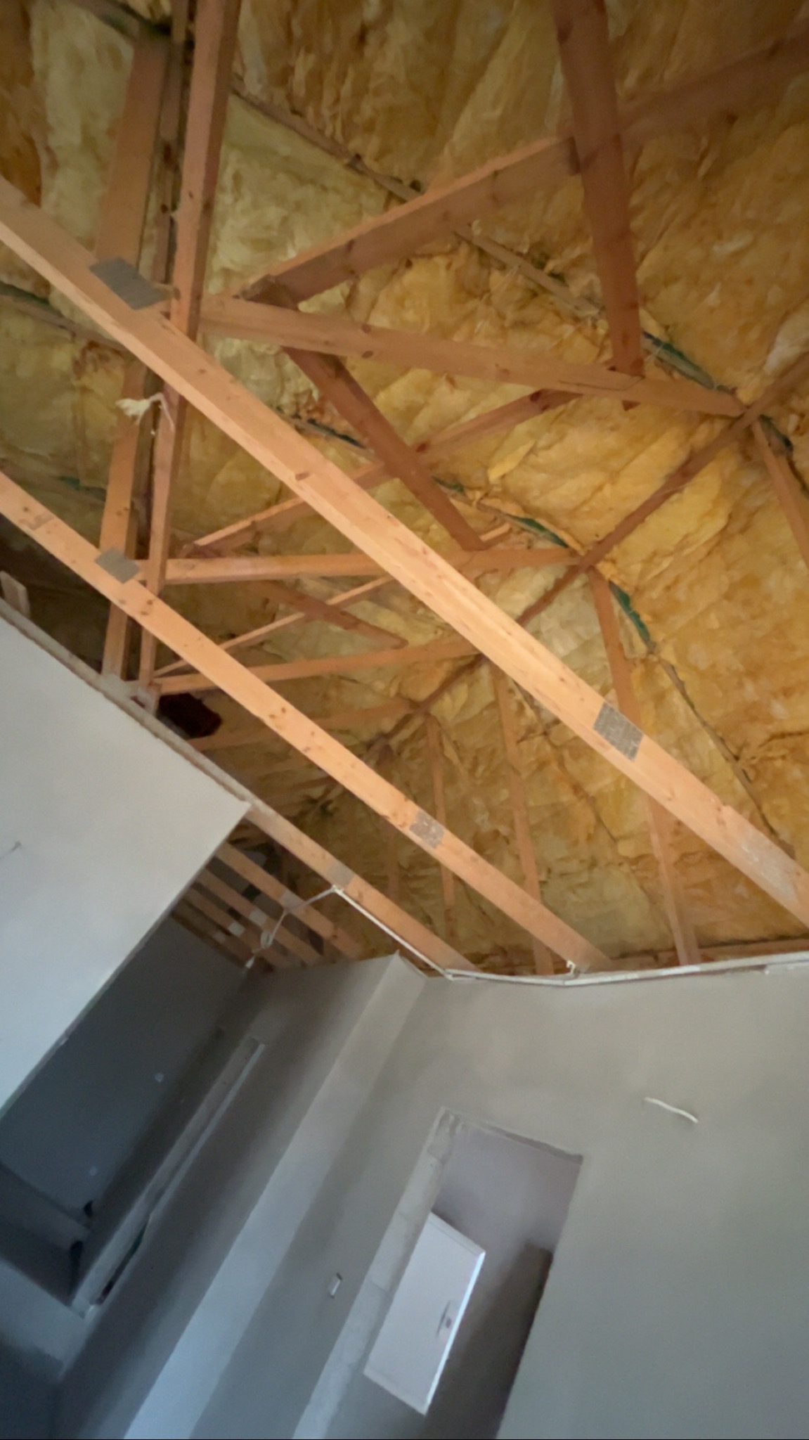 Ocieplone poddasze z widoczną konstrukcją drewnianą i izolacją z wełny mineralnej, fragment wykończonego wnętrza z szarą ścianą i białymi drzwiami.