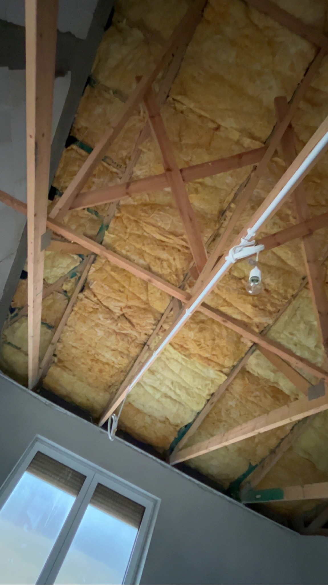 Poddasze z drewnianą konstrukcją i izolacją z wełny mineralnej, widoczne okno i instalacja elektryczna z żarówką. Ujęcie z dołu, ukośne.