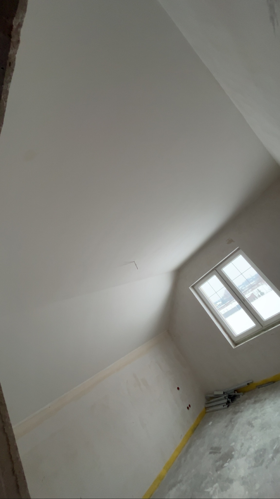 Poddasze po gładziowaniu, z oknem i widocznymi śladami prac wykończeniowych. Ściany i sufit w jasnym odcieniu, przygotowane do dalszych prac. Ujęcie z dołu, pod kątem.