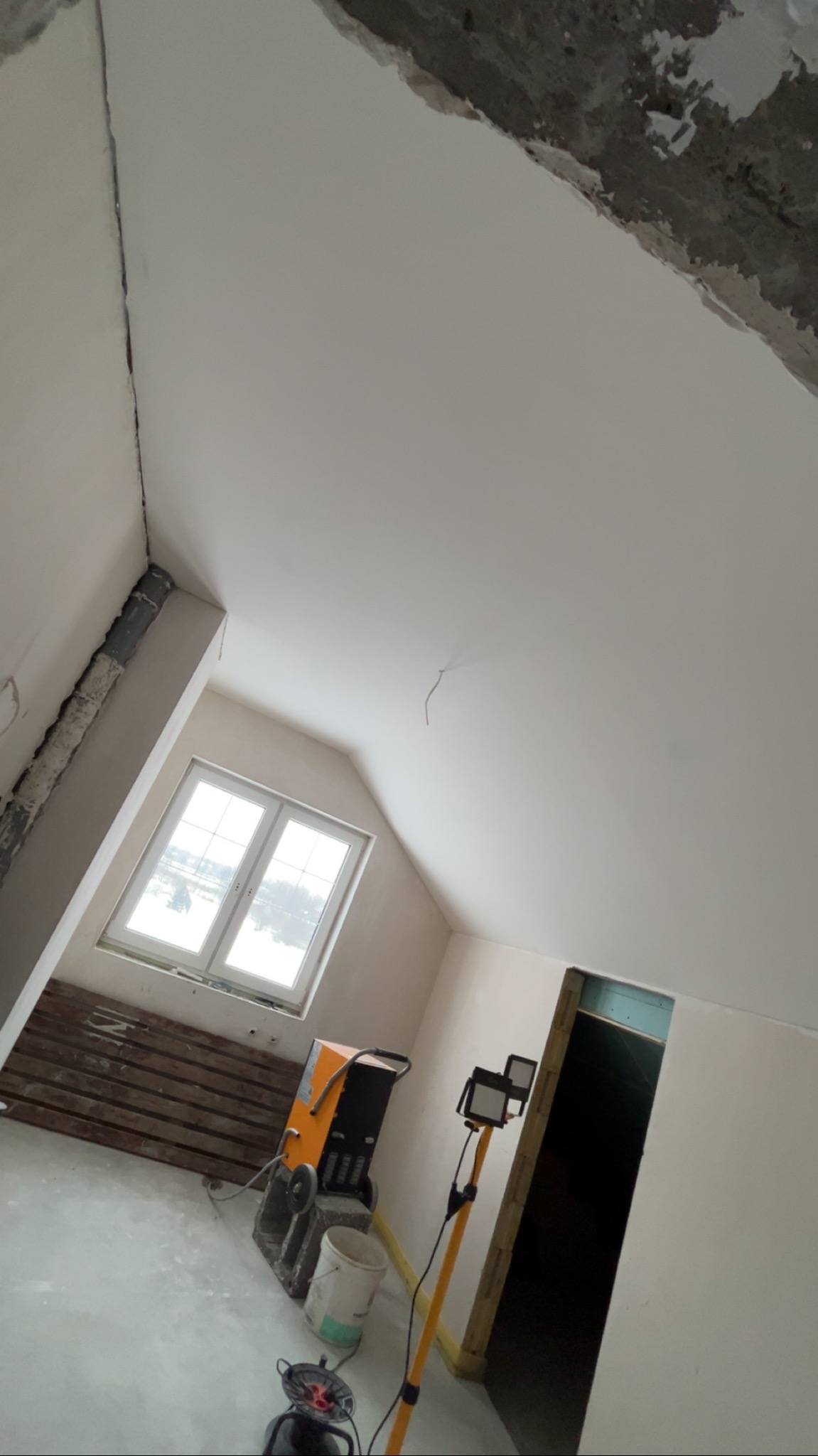 Poddasze po gładziowaniu, widoczne okno, lampa budowlana i osuszacz. Ściany i sufit świeżo pomalowane na biało. Prace wykończeniowe w trakcie realizacji.