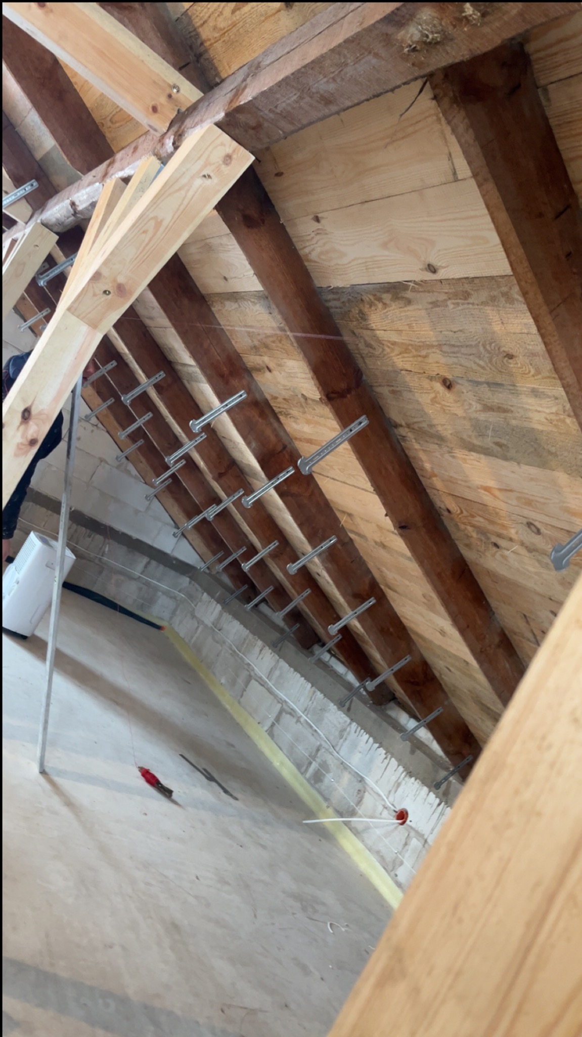 Poddasze w trakcie prac: drewniana konstrukcja dachu z zamocowanymi metalowymi elementami, widoczne przewody i narzędzia. Przygotowanie do ocieplenia.