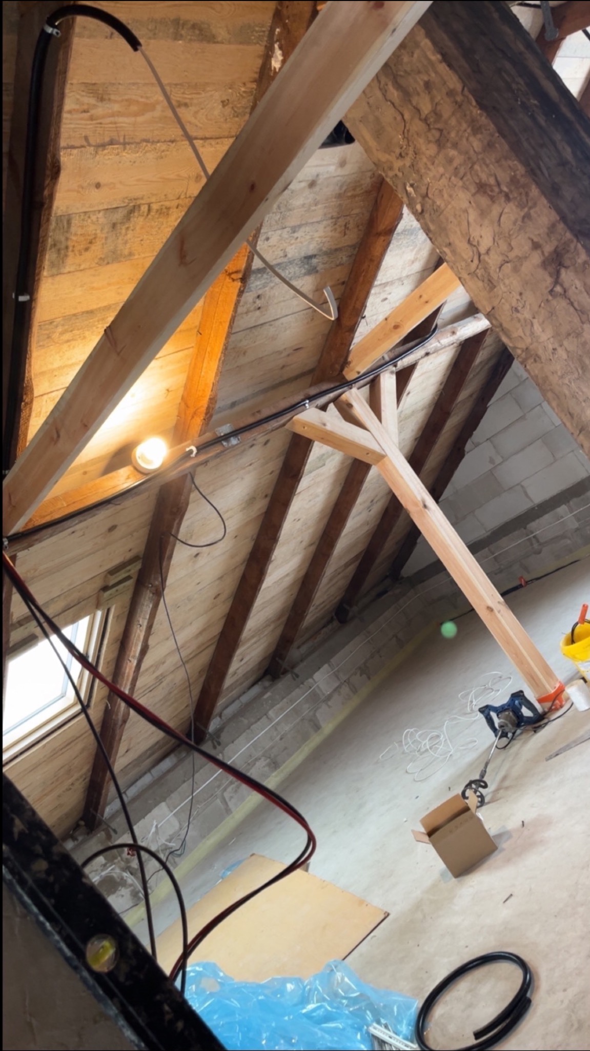 Skośny strych w trakcie prac: widoczne drewniane elementy konstrukcyjne, okno dachowe, instalacje elektryczne, narzędzia i materiały budowlane. Ujęcie z dołu.