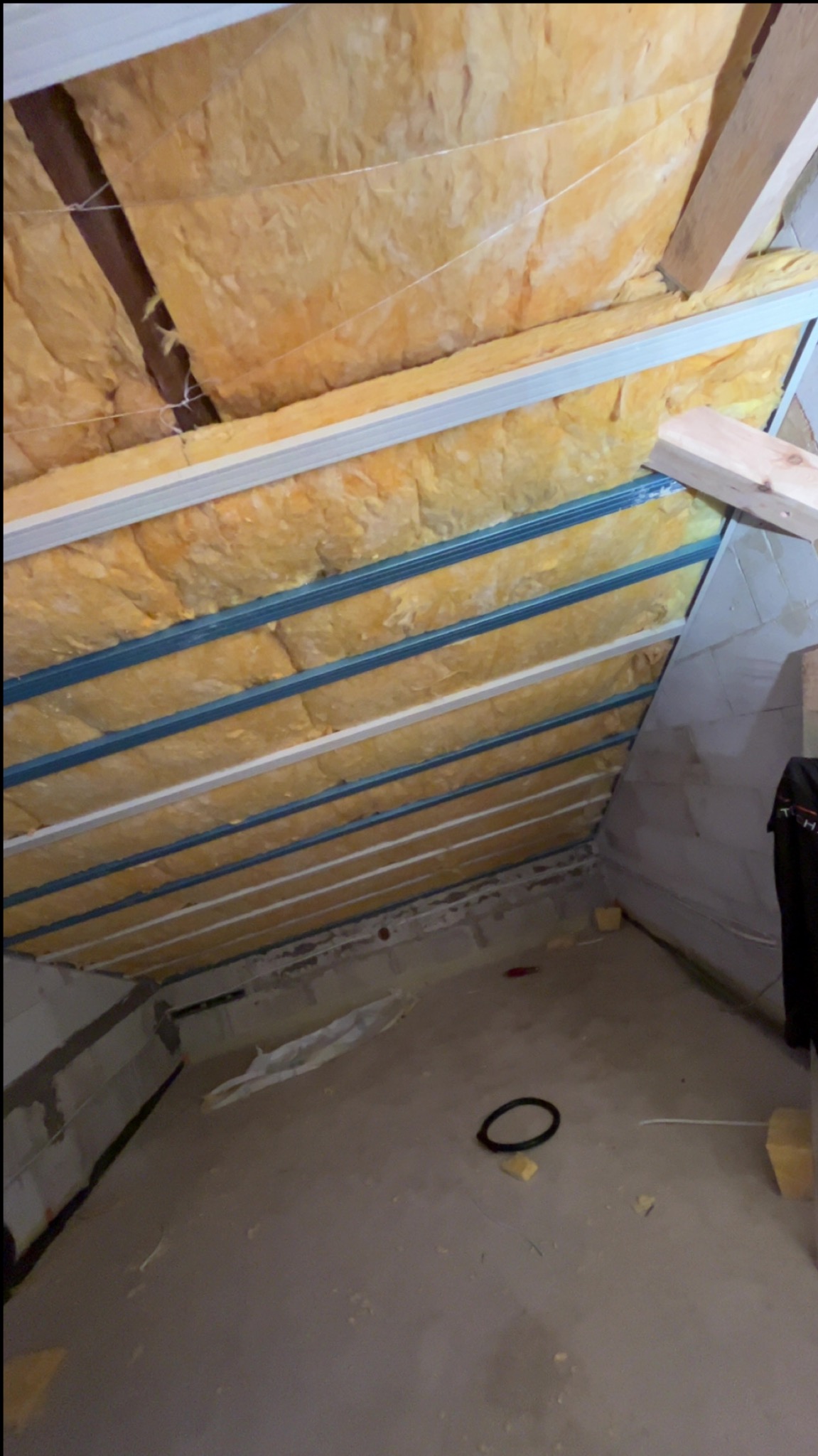 Skośne poddasze z ociepleniem z wełny mineralnej, widoczne metalowe profile konstrukcyjne i fragment betonowej ściany. Ujęcie z dołu, widać podłogę i elementy instalacji.