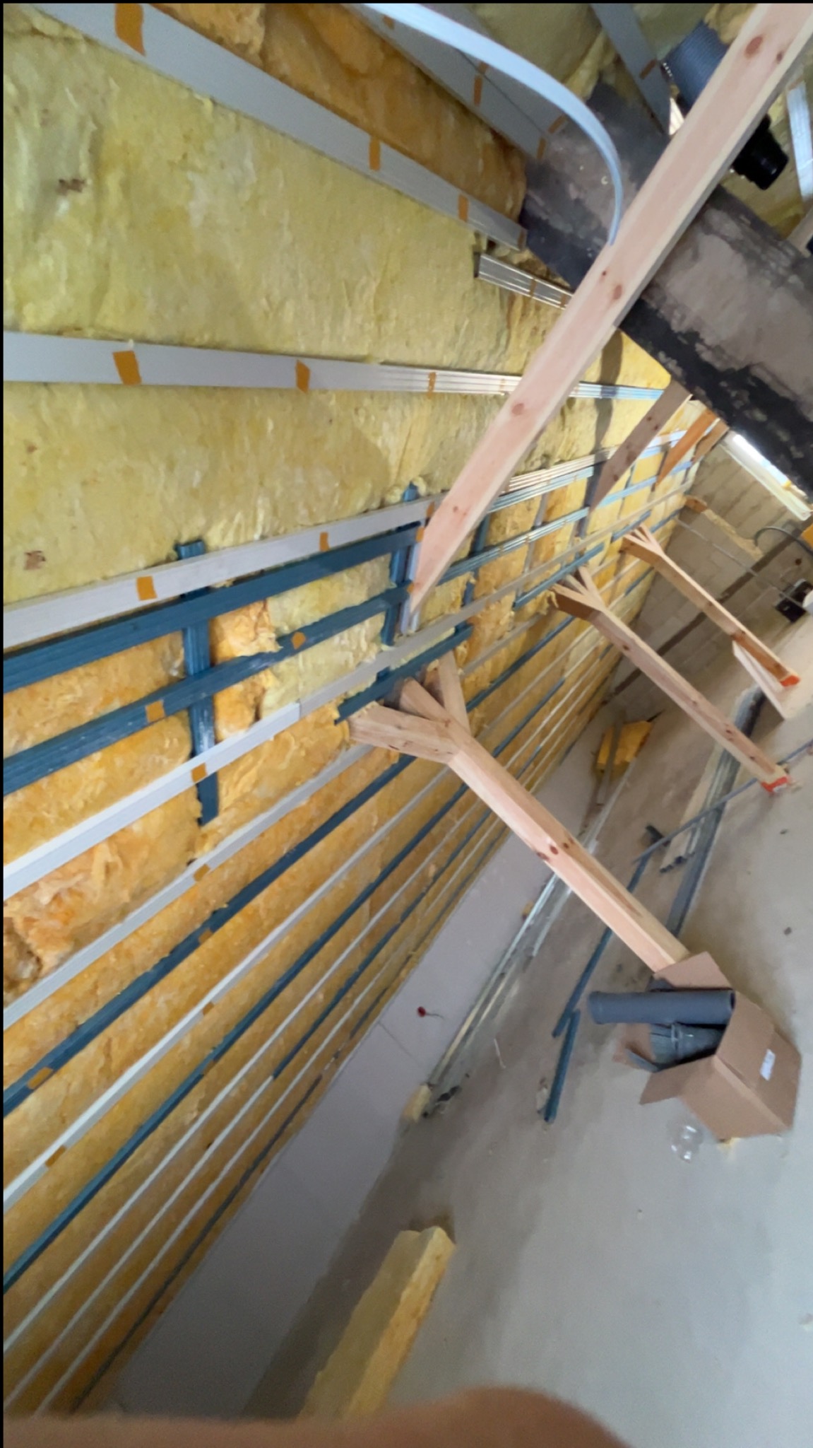 Skośny kadr na ocieplone wełną mineralną poddasze z widoczną konstrukcją drewnianą i metalowymi profilami. Fragment kartonu z rurami na podłodze.