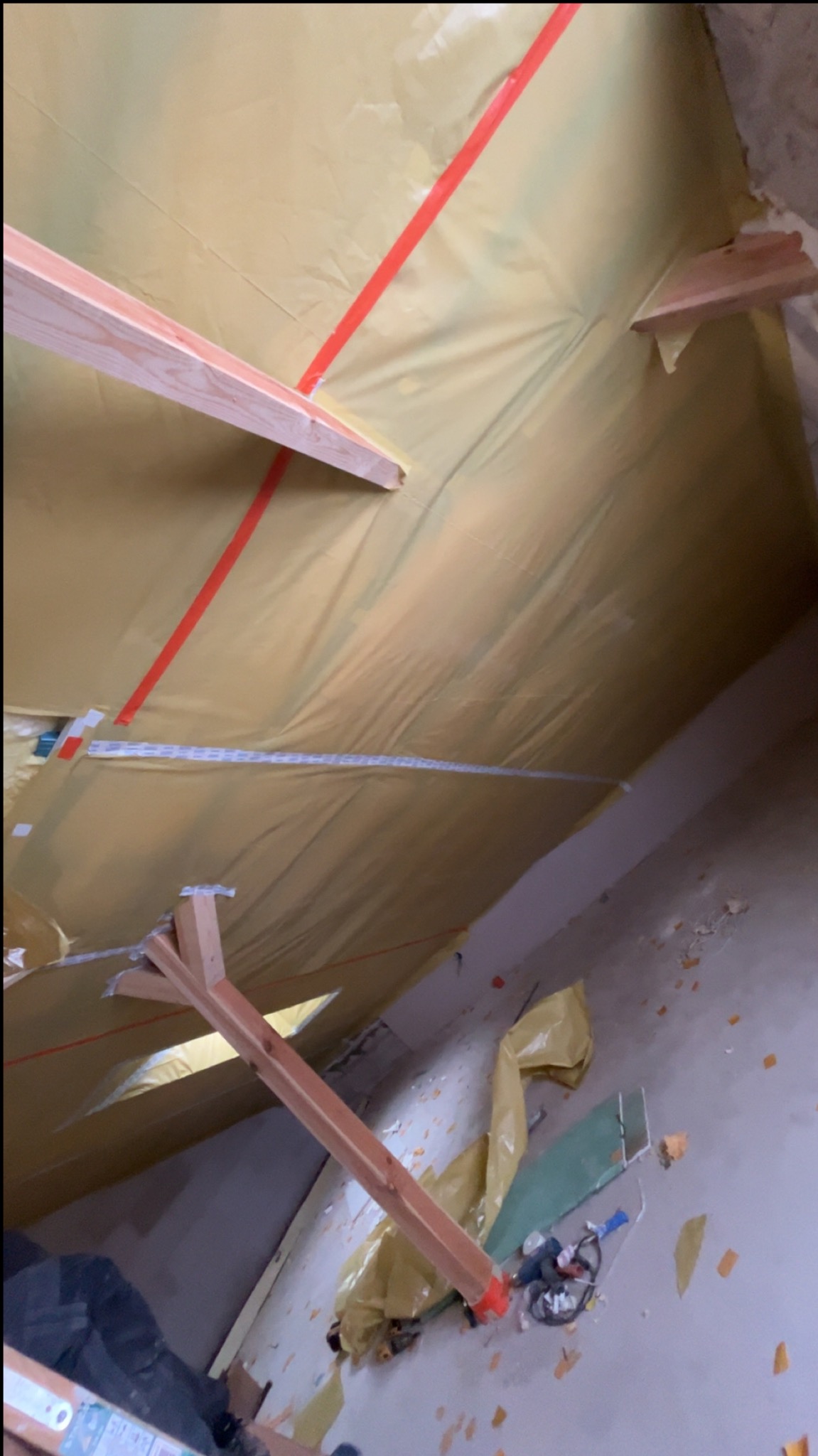 Żółta folia paroizolacyjna na poddaszu, mocowana do drewnianych belek czerwoną taśmą. Widoczne narzędzia i resztki materiałów na podłodze. Ujęcie z dołu.