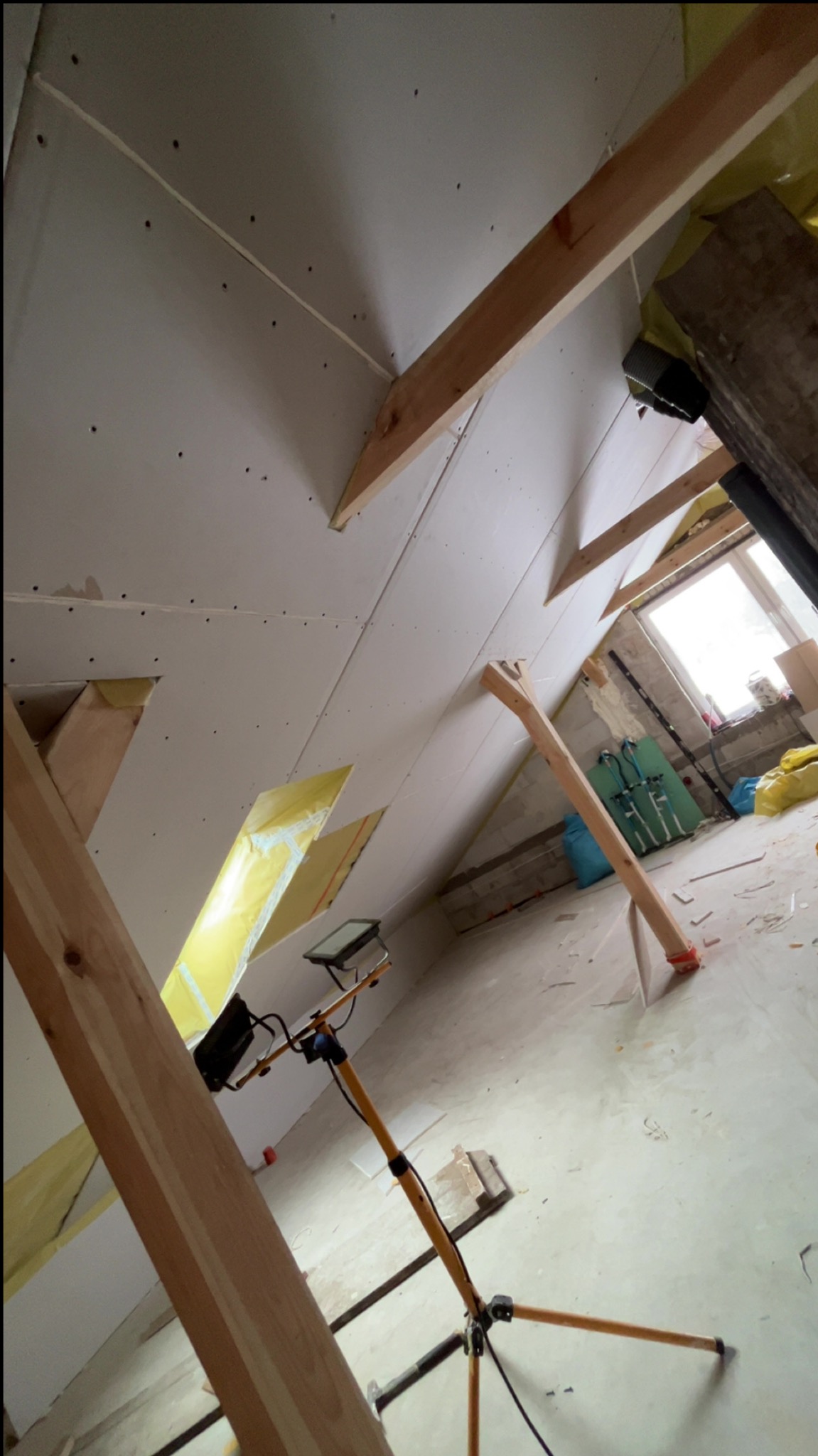 Skośny sufit poddasza z zamontowaną płytą gipsową, widoczne drewniane elementy konstrukcyjne i oświetlenie robocze. Prace wykończeniowe w trakcie realizacji.