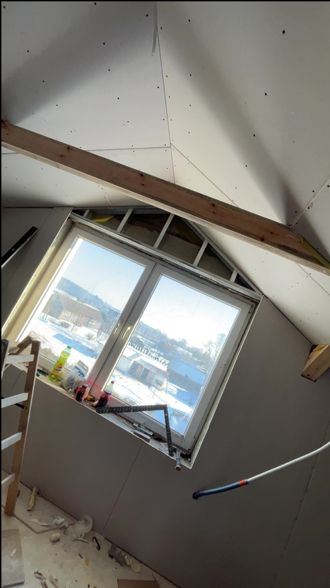 Poddasze z oknem dachowym w trakcie wykańczania płytami. Widoczne drewniane belki konstrukcyjne i narzędzia pracy. Prace wykończeniowe w jasnym wnętrzu.