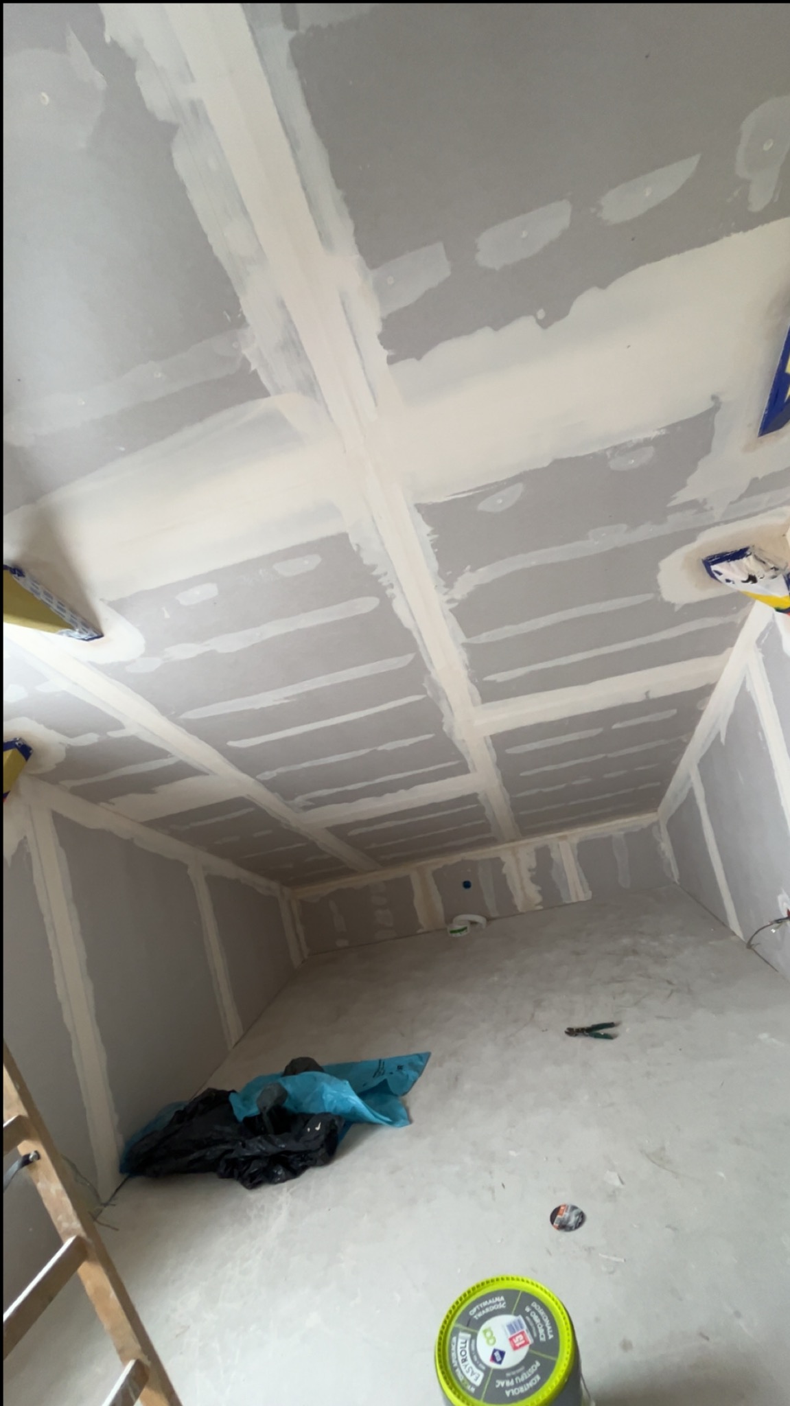 Płyty karton-gips na suficie i ścianach, wstępnie zaszpachlowane łączenia, widok z dołu, narzędzia na podłodze, przygotowanie do dalszych prac wykończeniowych.