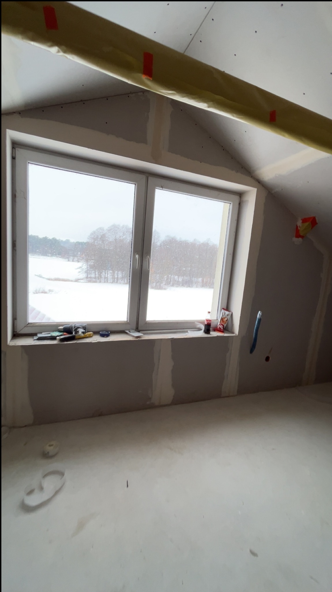 Poddasze z oknem po montażu płyt gipsowo-kartonowych, widoczne narzędzia na parapecie i taśma zabezpieczająca. Za oknem zimowy krajobraz.