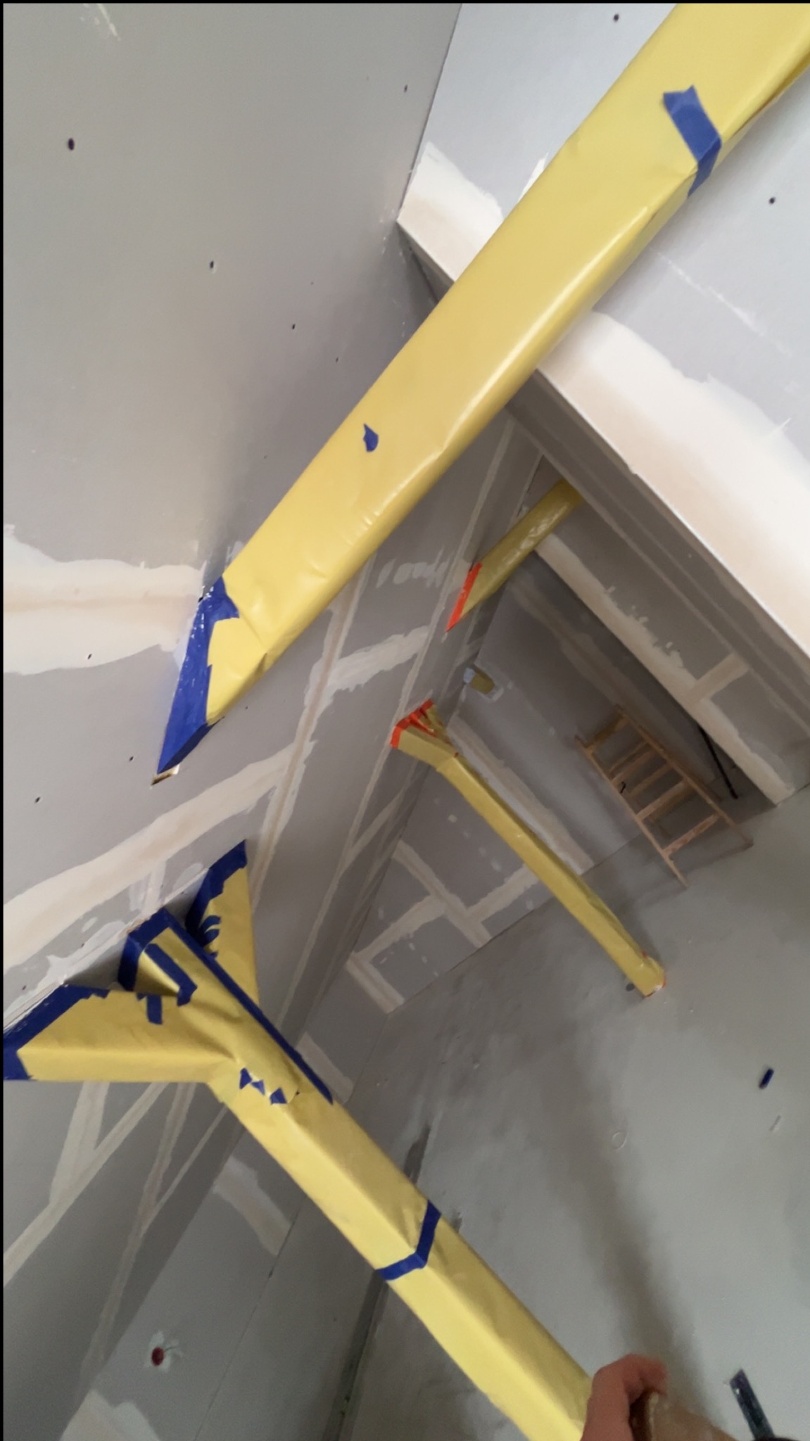 Skośne ujęcie fragmentu wnętrza w trakcie prac wykończeniowych; widoczne profile zabezpieczone folią i płyty kartonowo-gipsowe na ścianach.