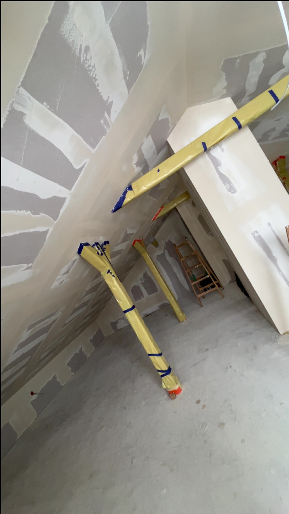 Skośny sufit i ściany po szpachlowaniu, zabezpieczone rury, drabina w tle. Prace wykończeniowe w pomieszczeniu o nieregularnym kształcie.