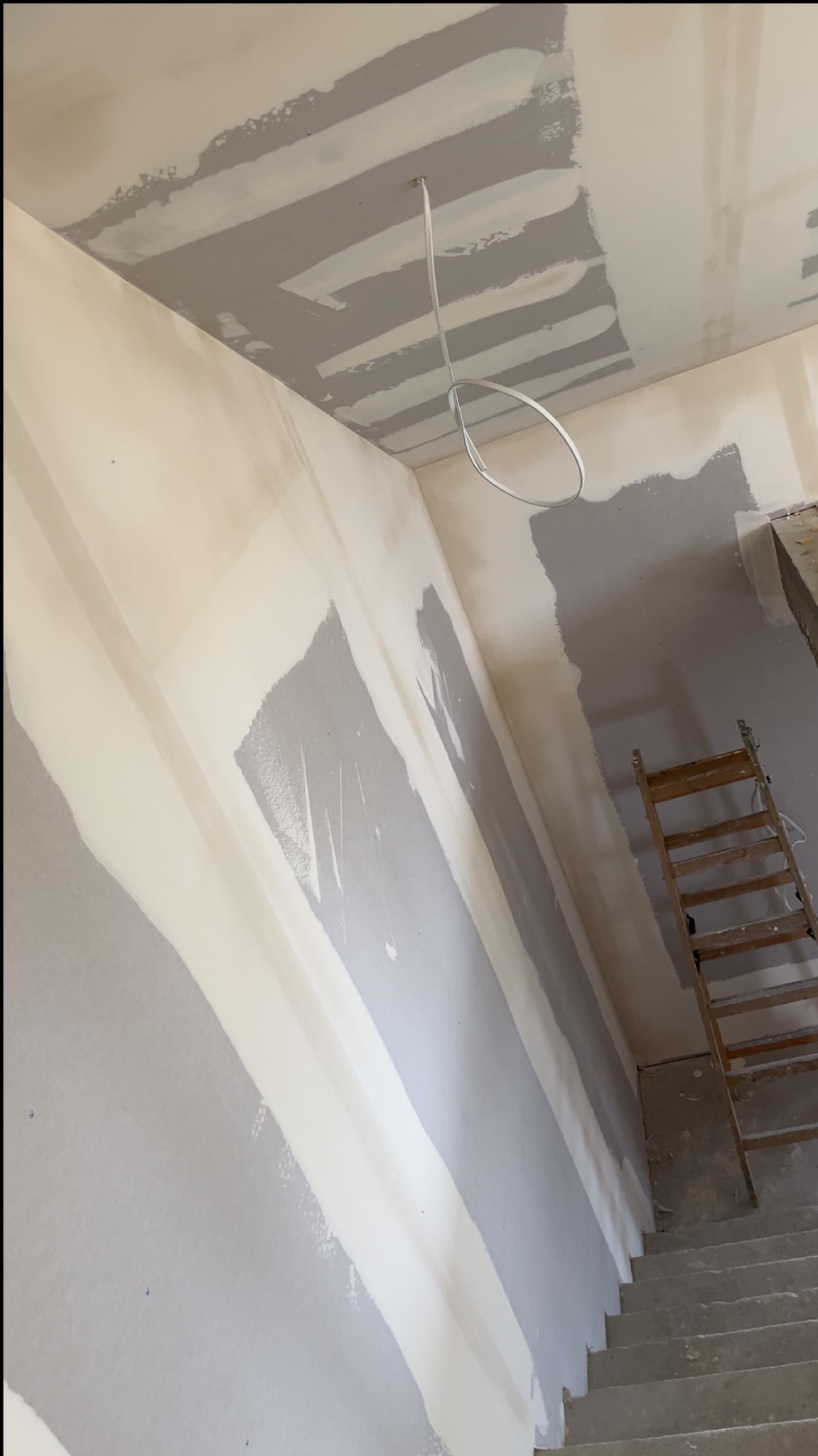 Ściana i sufit w trakcie szpachlowania, widoczne pasy gładzi, drabina oparta o ścianę. Wystający kabel elektryczny z sufitu. Ujęcie z dołu schodów.
