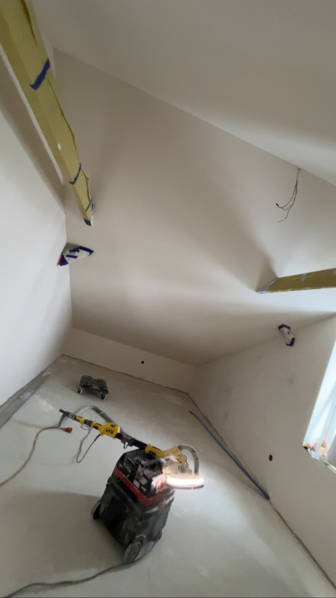 Szlifierka do gładzi na podłodze w pokoju ze skośnym sufitem po gipsowaniu. Widoczne taśmy malarskie zabezpieczające krawędzie. Przygotowanie do malowania.