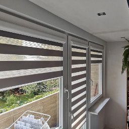 SOLAR - ŻALUZJE I ROLETY W KRAKOWIE JANUSZ SMOLEŃ - Zamontowane rolety dzień noc w kasecie, z brązowo-białymi pasami, widok na balkon i zieleń za oknem. Rolety zamontowane w trzech oknach.