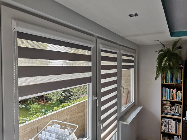 Zamontowane rolety dzień noc w kasecie, z brązowo-białymi pasami, widok na balkon i zieleń za oknem. Rolety zamontowane w trzech oknach.