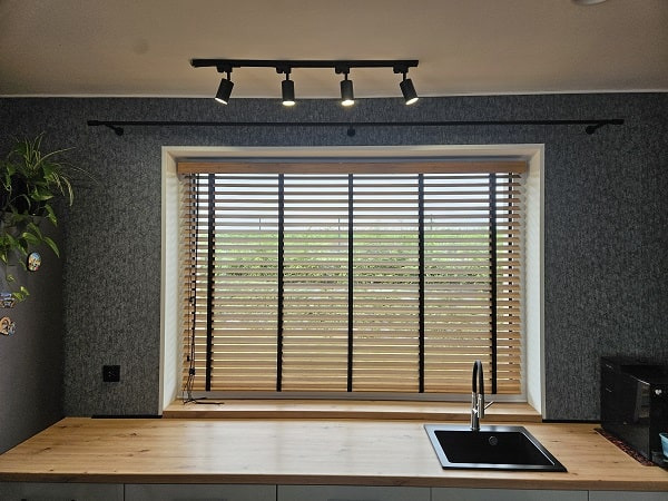 Kuchnia z drewnianymi żaluzjami 50mm przy oknie, widoczne czarne taśmy, szare ściany i oświetlenie punktowe. Naturalne światło dzienne.