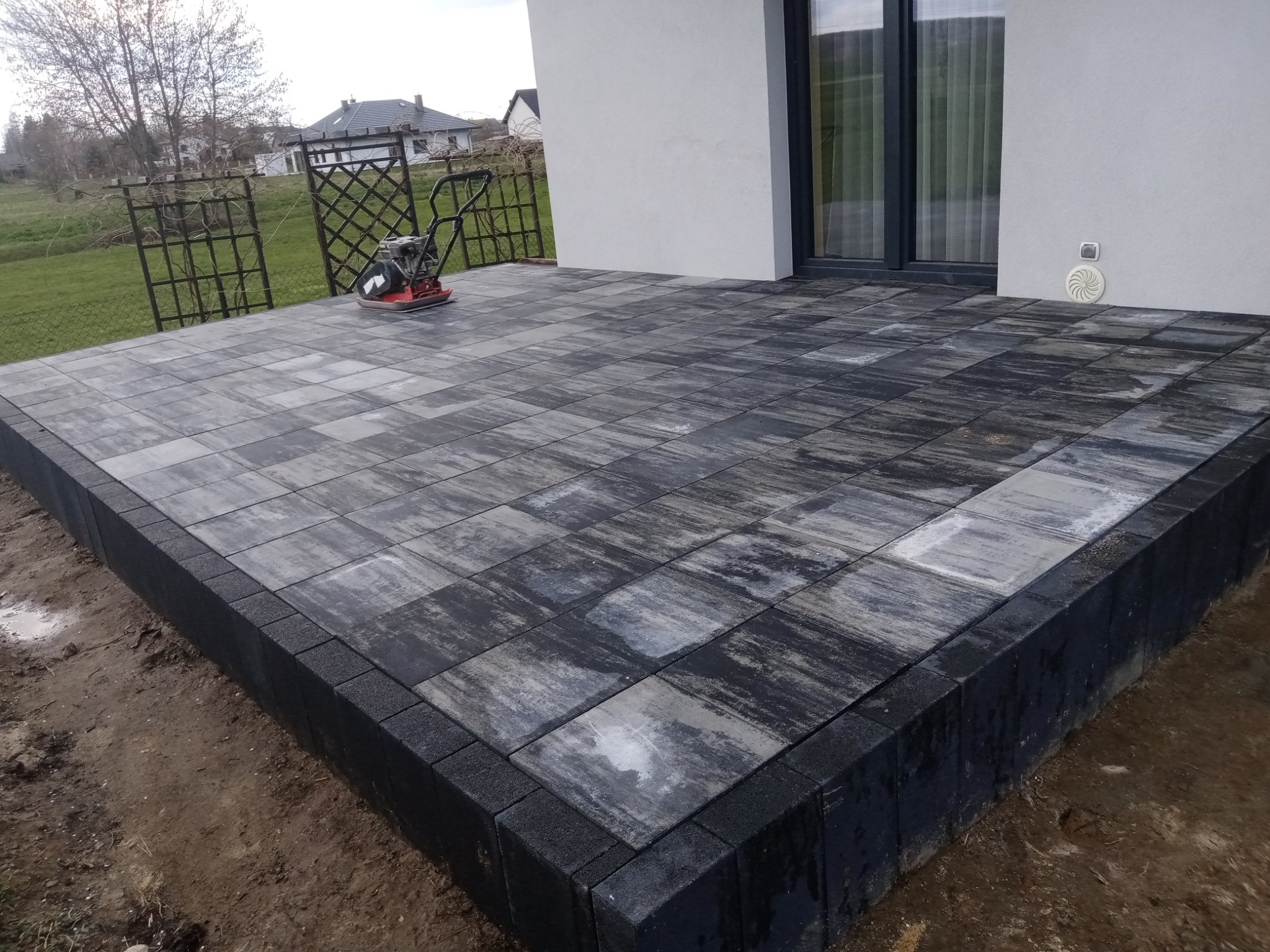 Nowo ułożony taras z kostki brukowej w odcieniach szarości i czerni, z widoczną zagęszczarką w tle, na tle trawnika i nowoczesnego domu z dużym oknem.