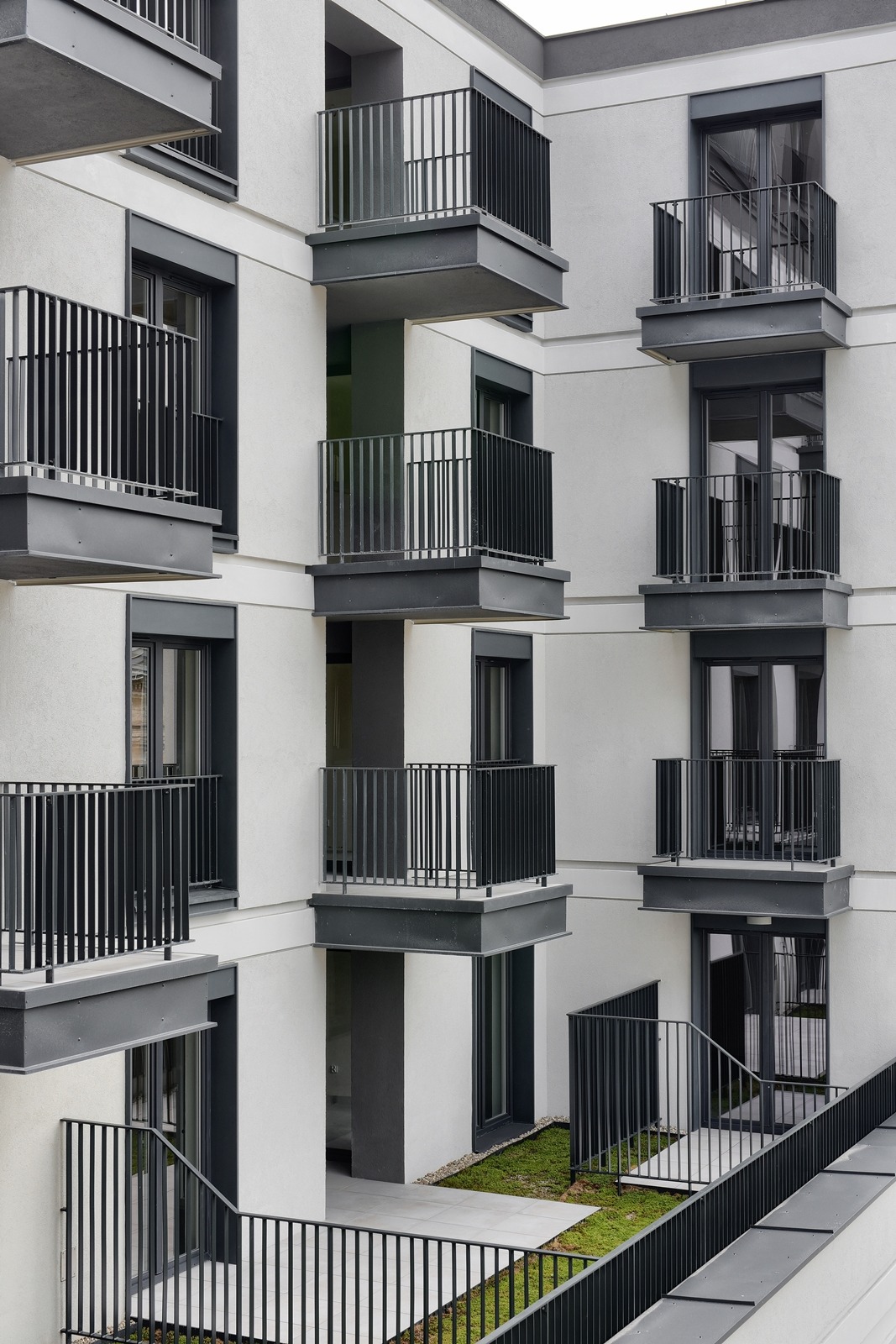 Fasada nowoczesnego budynku mieszkalnego z balkonami o stalowych balustradach i oknami w grafitowych ramach, z fragmentem zielonego dachu na parterze.