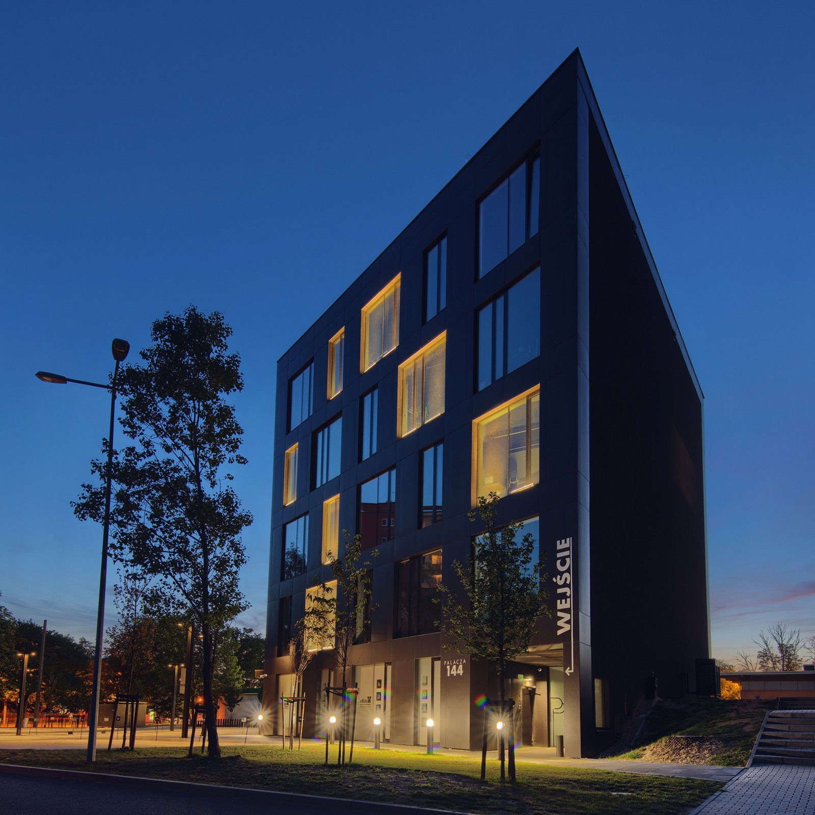 Nowoczesny, czarny budynek biurowy z podświetlonymi oknami i napisem 'Wejście' w Poznaniu, sfotografowany o zmierzchu, z widocznymi drzewami i lampami ulicznymi.