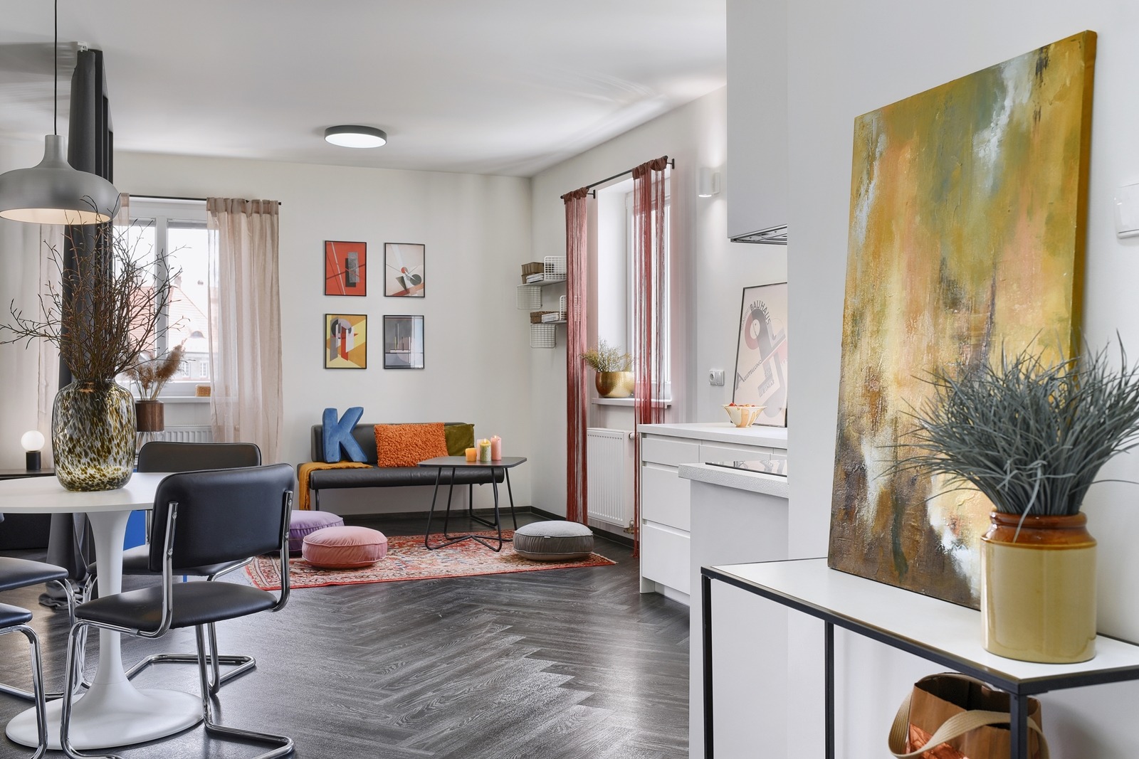 Jasne, przestronne wnętrze mieszkania z parkietem w jodełkę, okrągłym stołem z bukietem w wazonie, sofą z dekoracyjnymi poduszkami i abstrakcyjnym obrazem na ścianie, utrzymane w neutralnej...