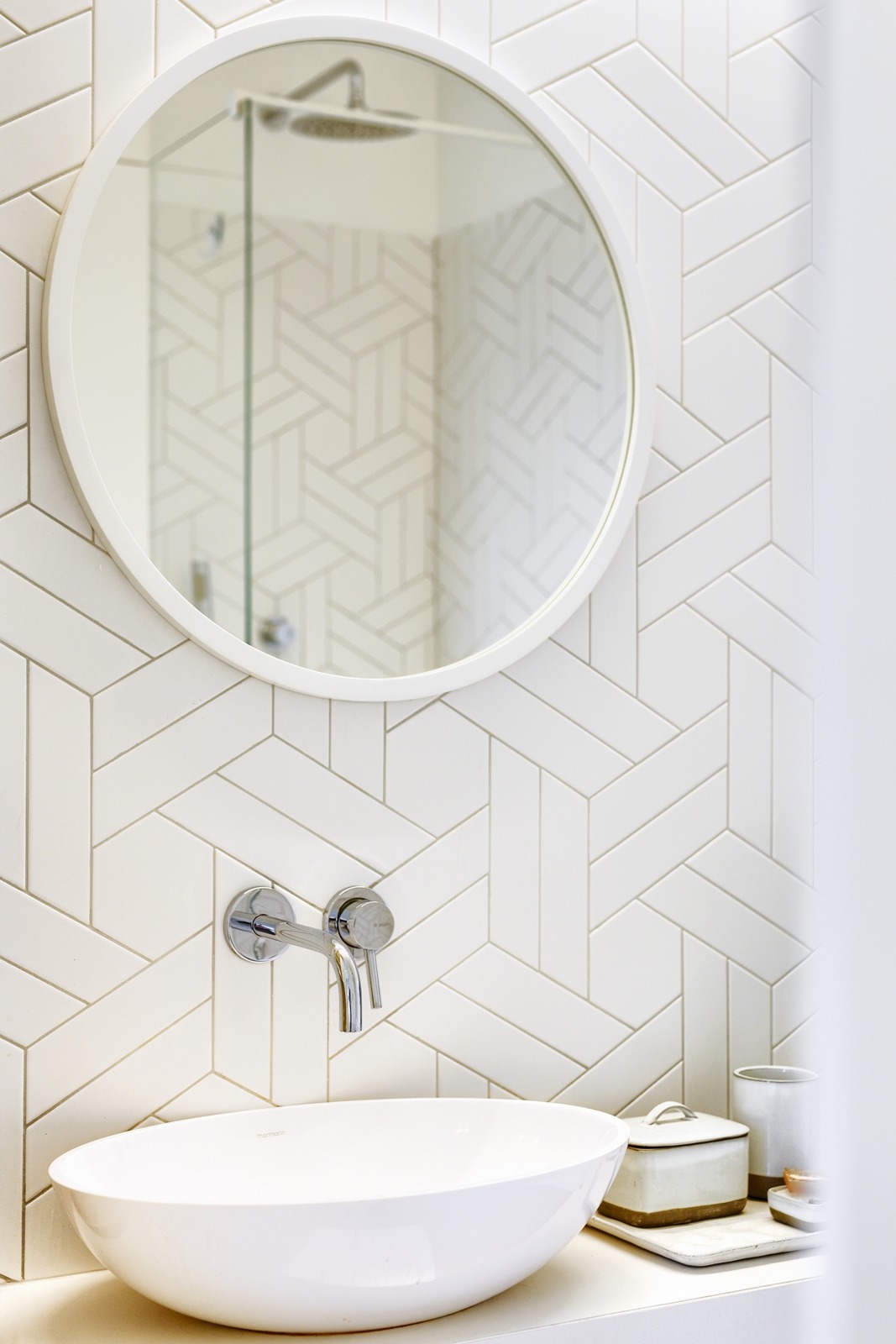 Nowoczesna łazienka z białą umywalką nablatową, chromowaną armaturą, okrągłym lustrem i geometrycznymi płytkami na ścianie, odbicie prysznica w lustrze.