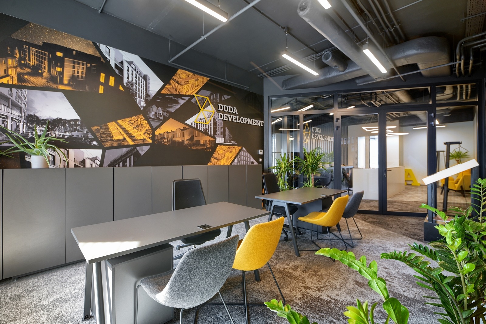 Nowoczesne biuro z szarymi biurkami, kolorowymi krzesłami i graficzną ścianą z logo 'DUDA Development' w geometryczne wzory przedstawiające architekturę.