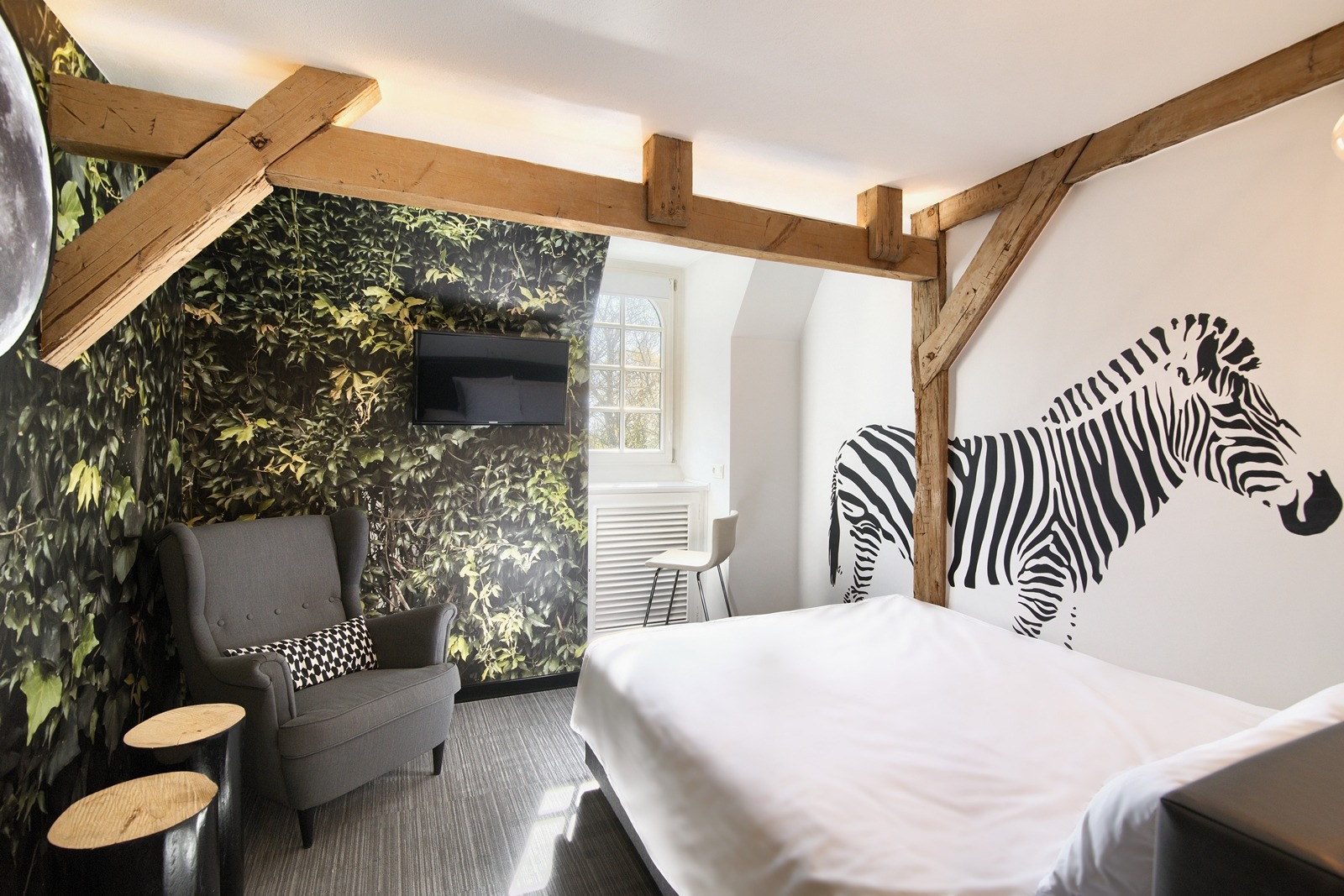 Pokój hotelowy z tapetą w liście, ścianą z motywem zebry, drewnianymi belkami stropowymi, szarym fotelem i stolikami pomocniczymi.
