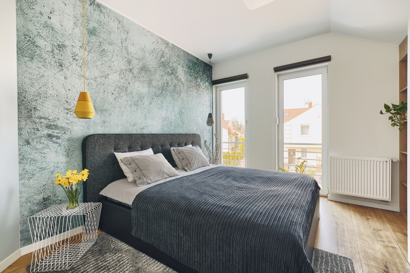 Sypialnia z łóżkiem z szarą tapicerką, dekoracyjną ścianą w odcieniach zieleni i żółtą lampą wiszącą, na stoliku nocnym wazon z żółtymi narcyzami.